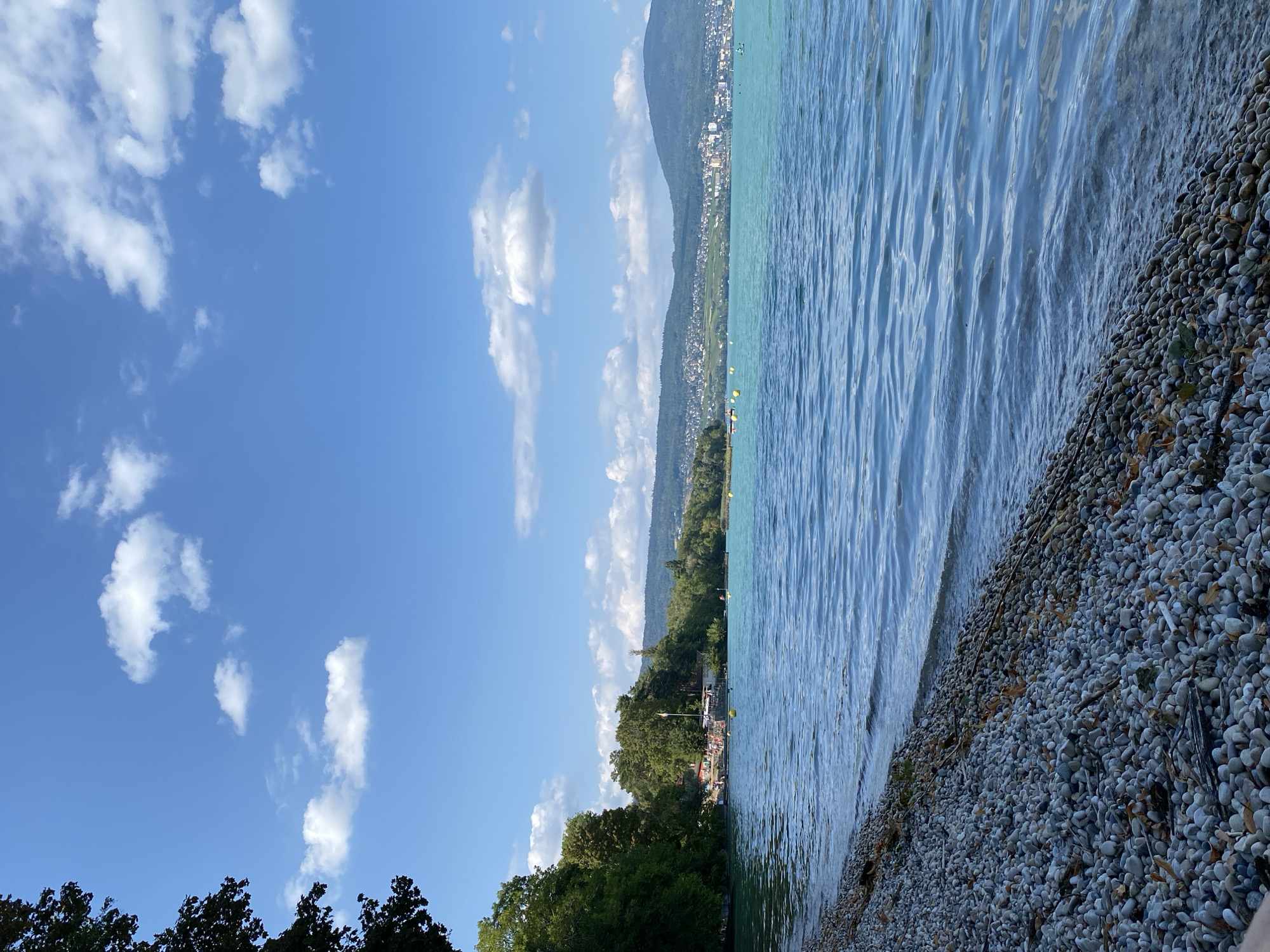A serene lakefront scene unfolds under a bright blue sky dotted with scattered white clouds. The foreground is dominated by a pebble-strewn shore, where small, smooth stones of varying shades of gray and brown lie at the edge of the tranquil, blue-green water. Gentle ripples disturb the lake's surface, reflecting the sky above.
To the left, a dense cluster of lush green trees creates a natural border along the water's edge. Beyond the trees, on the lake, a few yellow buoys are visible, marking a designated area. In the middle ground, a small beach area is evident, with several people scattered along the shore and in the shallow water, suggesting a recreational setting. Buildings with red roofs are nestled amongst the trees, and a tall flagpole stands prominently.
The background is a picturesque panorama of rolling hills covered in greenery and dotted with buildings, extending towards the horizon. The city appears to climb the slopes of the hills, with more distant mountains forming a soft, hazy backdrop. The overall impression is one of a peaceful, sunny day at a popular lakeside spot. This scene is consistent with the natural beauty and recreational opportunities found in the vicinity of Cortaillod, Switzerland, a commune situated on the shores of Lake Neuchâtel.