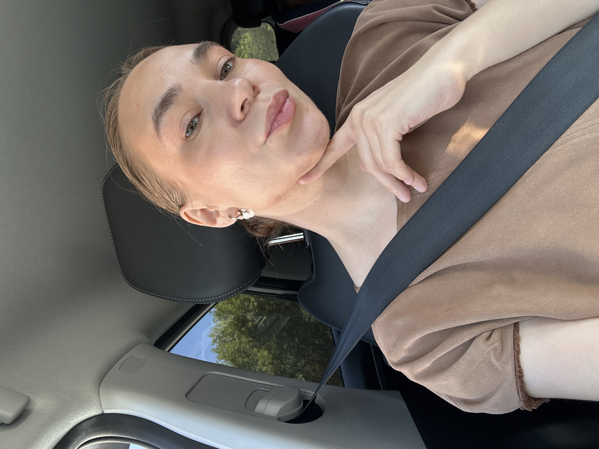 A light-skinned woman with brown hair pulled back into a bun is posing for a selfie while sitting in a car. She wears a brown top and a seatbelt. Her right hand is positioned with her index finger touching her chin. The background visible through the car window shows trees, suggesting an outdoor setting. Given the mention of Chaikovsky, Russia, the photo was likely taken in or near this city, although no specific location is discernible from the image alone.