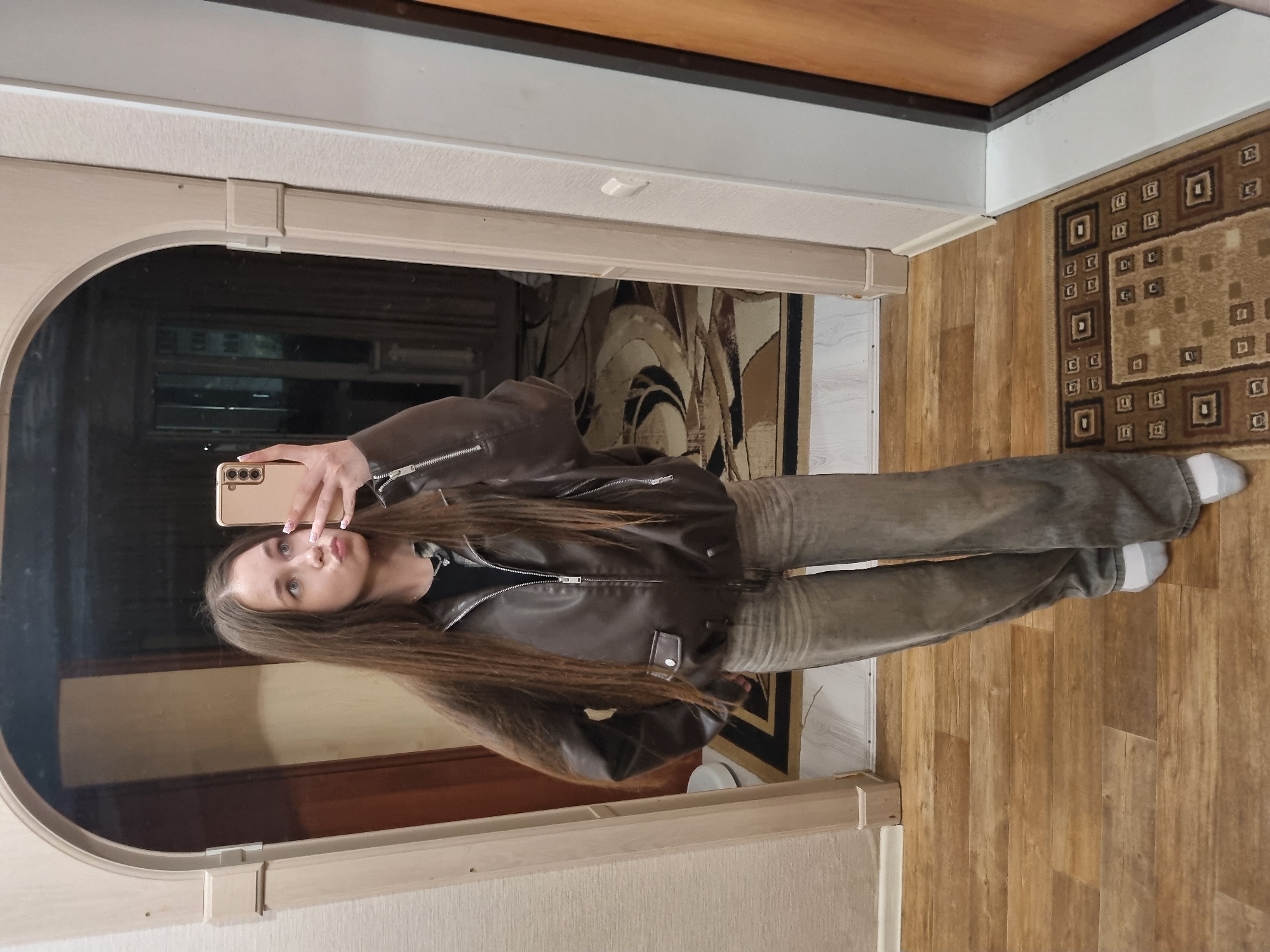 A young woman stands in a room taking a selfie in a mirror. She wears a brown leather jacket, gray jeans, and gray and white socks. The room has hardwood flooring, a patterned rug, and light-colored walls. The mirror is framed with a white or light-colored frame. The image is taken indoors. The location is not discernible from the image, therefore I cannot say it is Chaikovsky, Russia.