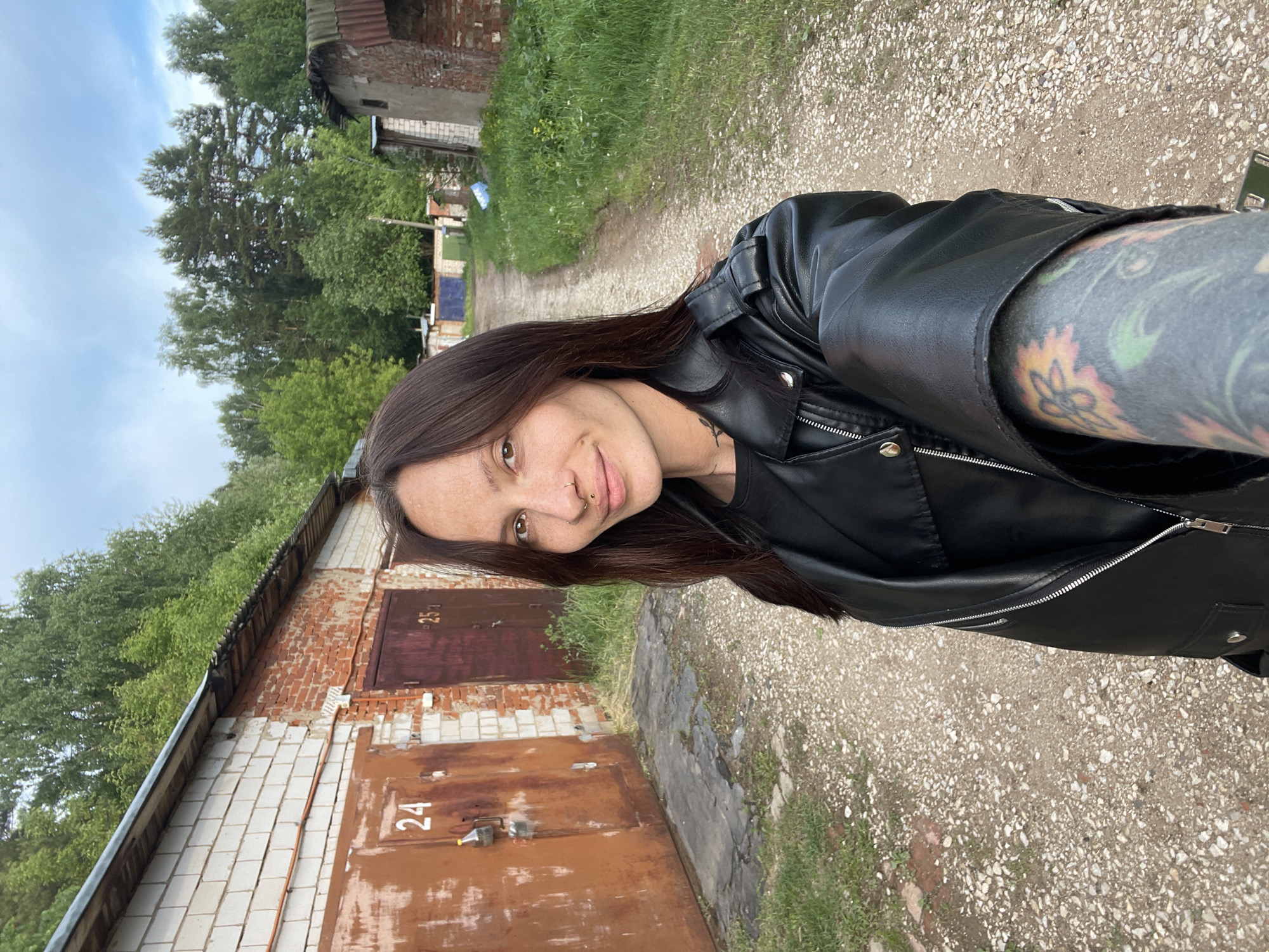 A woman stands on a gravel-covered path next to a row of brick garages. She is wearing a black leather jacket and has a tattoo on her arm. The garages are numbered 24 and 25. The location is in a rural area, with trees and greenery visible in the background. The sky is overcast. The woman has dark hair and a nose ring. The image appears to be a selfie. The city of Oktyabr'skii is not evident.
