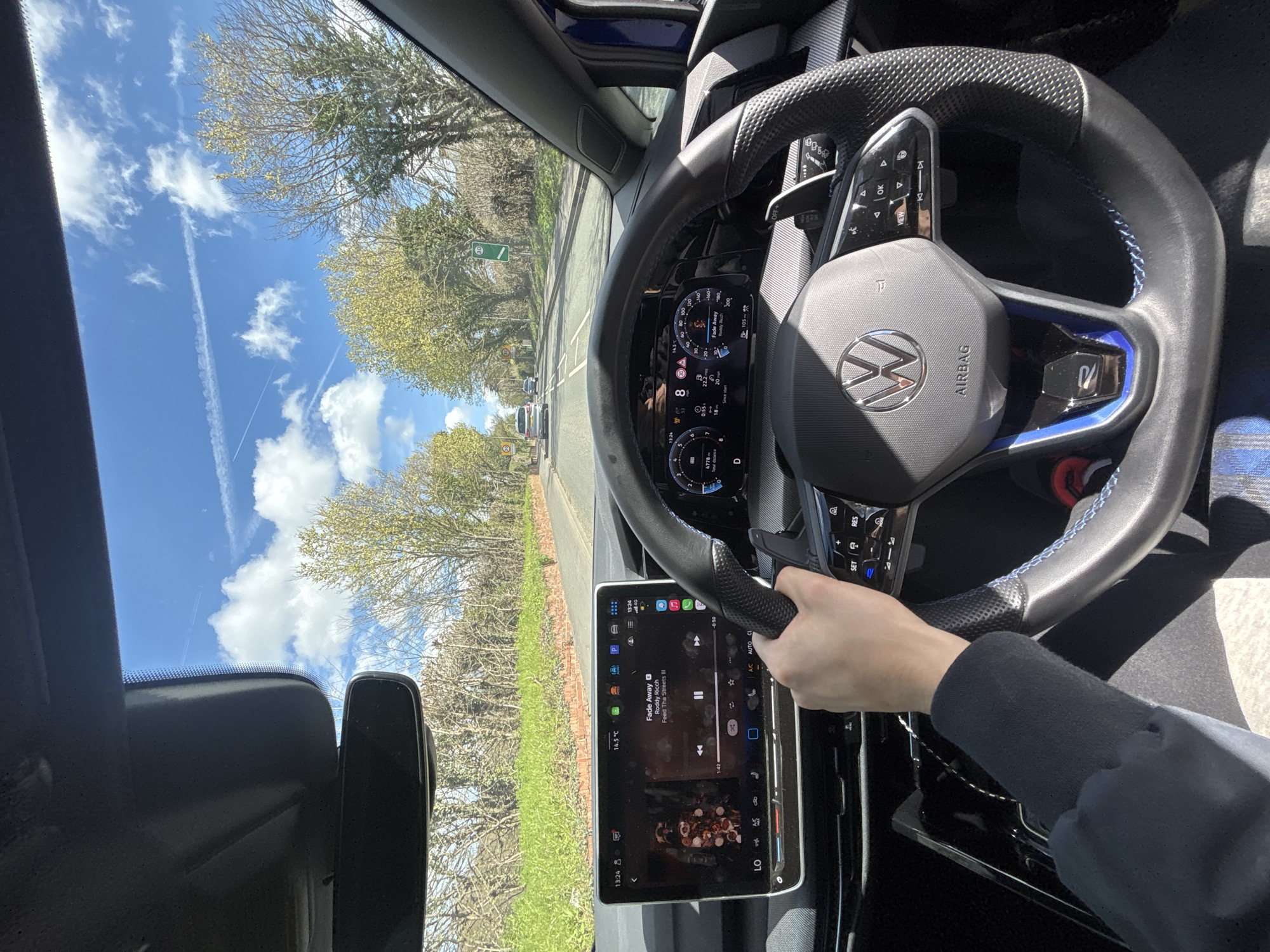 A photo, taken from the driver’s seat of a car, shows a person driving on a tree-lined road under a blue sky with contrails. The vehicle’s dashboard features a steering wheel with a Volkswagen logo, a digital speedometer, and a large infotainment screen displaying music information. The screen shows “Fade Away” by Roddy Ricch is playing. The speedometer reads 8 mph, and the dashboard shows 14.5 °C (58.1 °F). The photo may have been taken in or near Broxbourne, United Kingdom, given that a green road sign is partially visible in the background, which are commonly found in the UK. The location cannot be verified definitively with the available information.