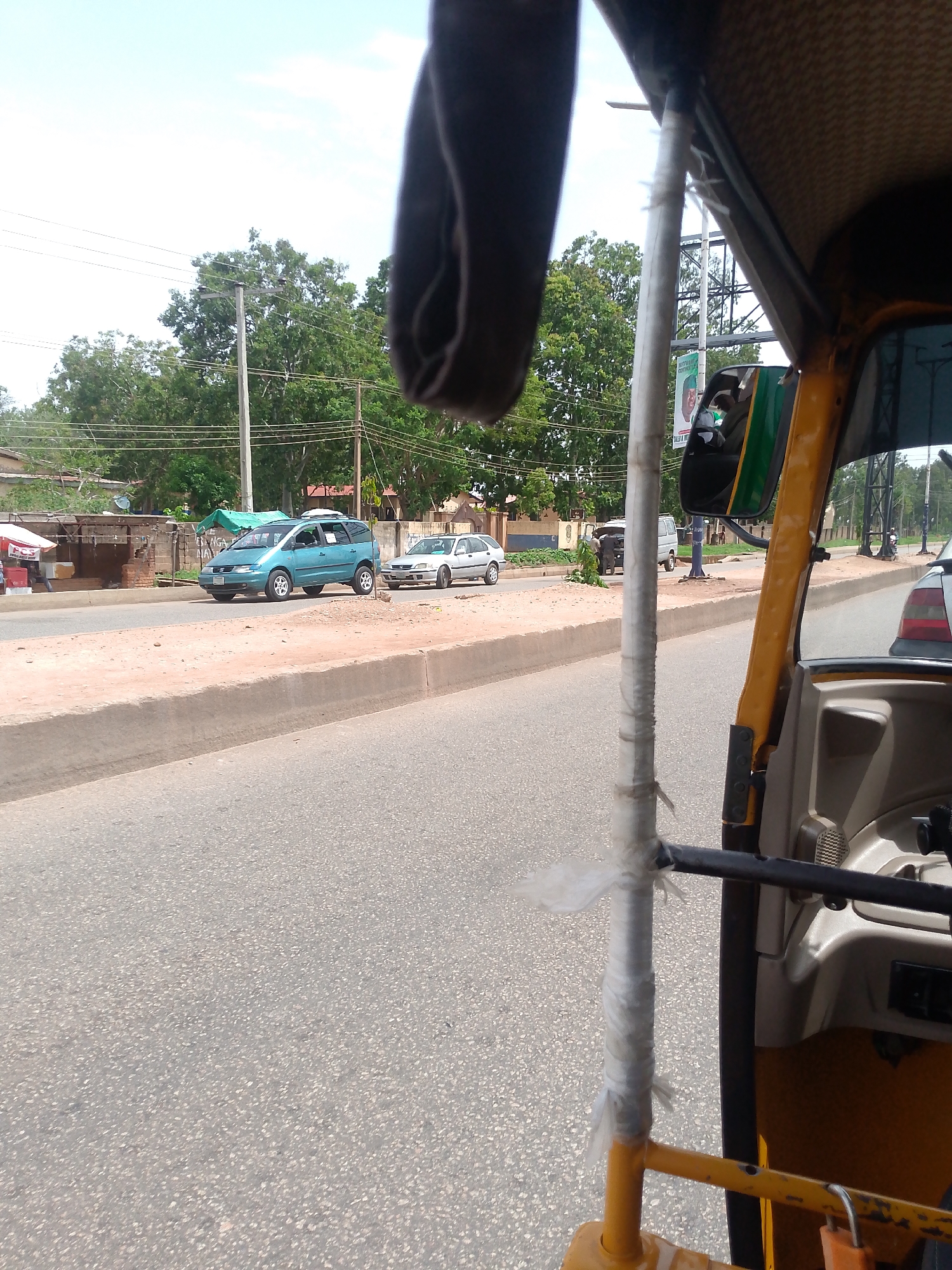The image depicts a street scene from inside a yellow tuk-tuk in Bukuru, Nigeria. A blue minivan and a silver sedan are visible on the opposite side of the road. Other vehicles are further down the road. The road is paved and has a raised median. Trees and buildings are visible in the background. Power lines stretch across the sky. A political poster is attached to a utility pole. The tuk-tuk's interior is visible, including the steering mechanism wrapped with a white material.