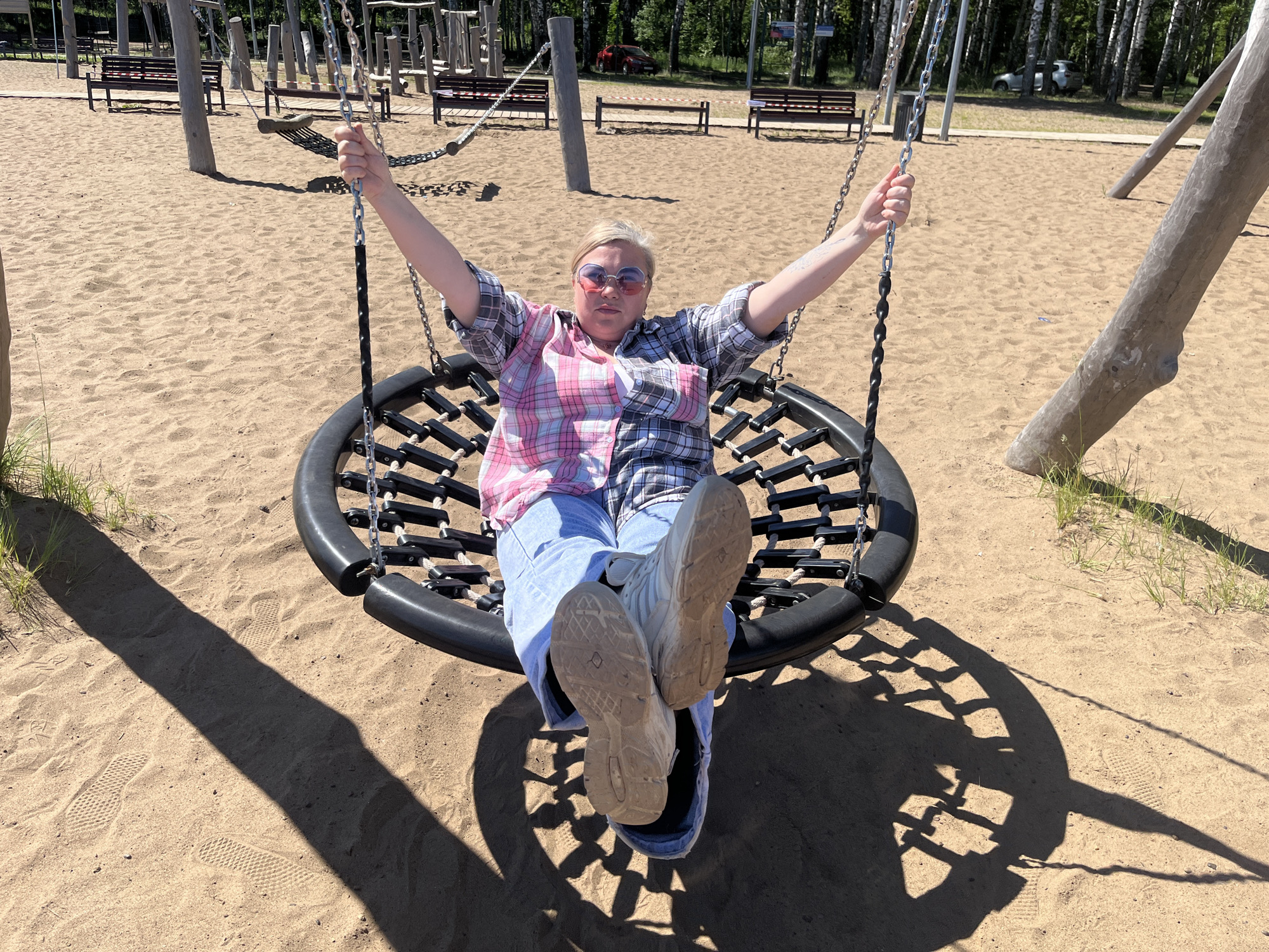The image shows a woman sitting on a round swing in a sandy playground. She is wearing a plaid shirt, blue jeans, and white sneakers. Her arms are outstretched, and she is looking at the camera. The swing is made of black rope and has a black frame. In the background, there are trees, benches, and other playground equipment. The ground is covered in sand, with footprints visible. The lighting suggests it is a sunny day. The location is identified as a playground in Galewo, Russia.