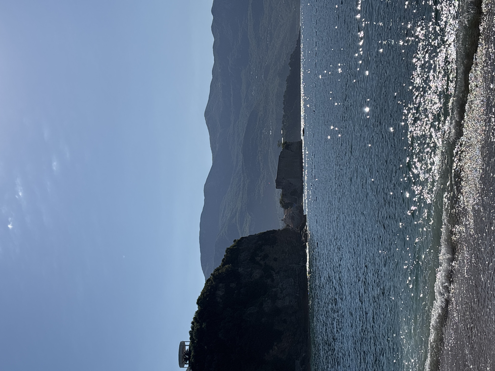 This is a vertical, medium-shot photograph of a coastline with mountains in the background. The sky is clear and blue with a few faint clouds. The ocean is a deep blue and sparkles with sunlight. Gentle waves lap at a sandy beach in the foreground. On the left side of the image, a dark, tree-covered cliff juts out into the sea, topped with a circular structure. In the middle ground, a stone fortress sits on a rocky outcrop. The background is filled with rolling, green mountains. The lighting suggests a sunny day, likely in the late morning or early afternoon.
