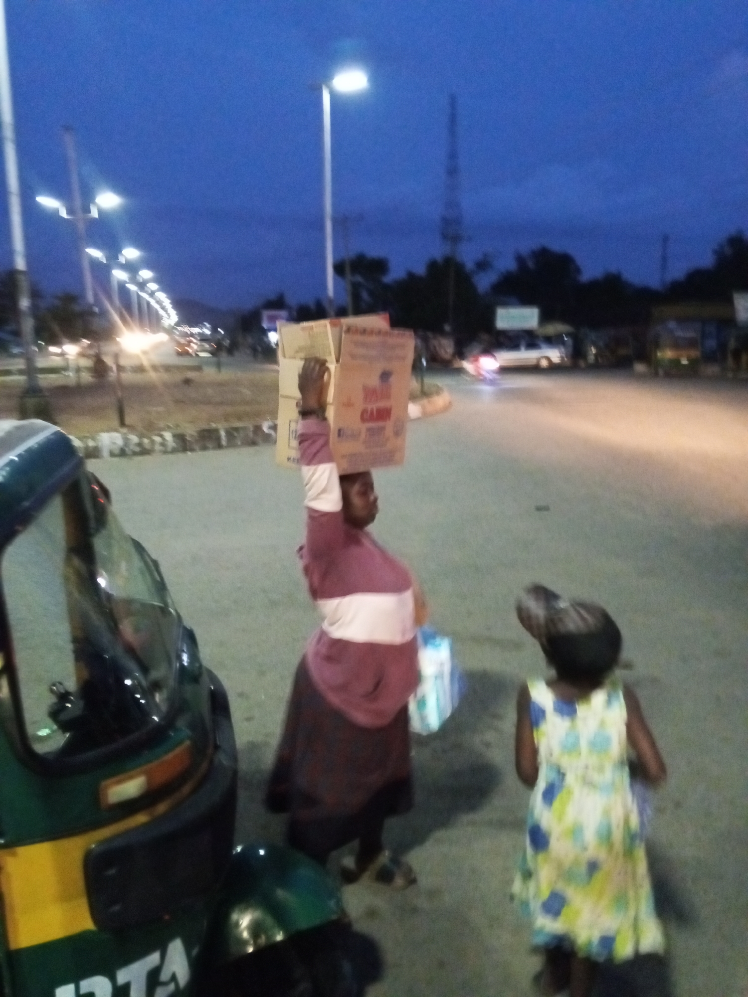 In Bukuru, Nigeria, during the twilight hours, a woman balances a large cardboard box labeled "CANON" on her head. She wears a maroon and white horizontally striped top and a dark skirt. Beside her, a young girl in a light-colored dress with a floral pattern of blue and yellow is visible from behind, also appearing to carry something. To the left, a green and yellow auto-rickshaw, a common mode of transportation in Nigeria, is parked. The scene unfolds on a paved road, illuminated by streetlights and the headlights of passing vehicles. In the background, a street lined with lights stretches into the distance, flanked by trees and the silhouettes of distant structures, suggesting an urban or peri-urban environment.