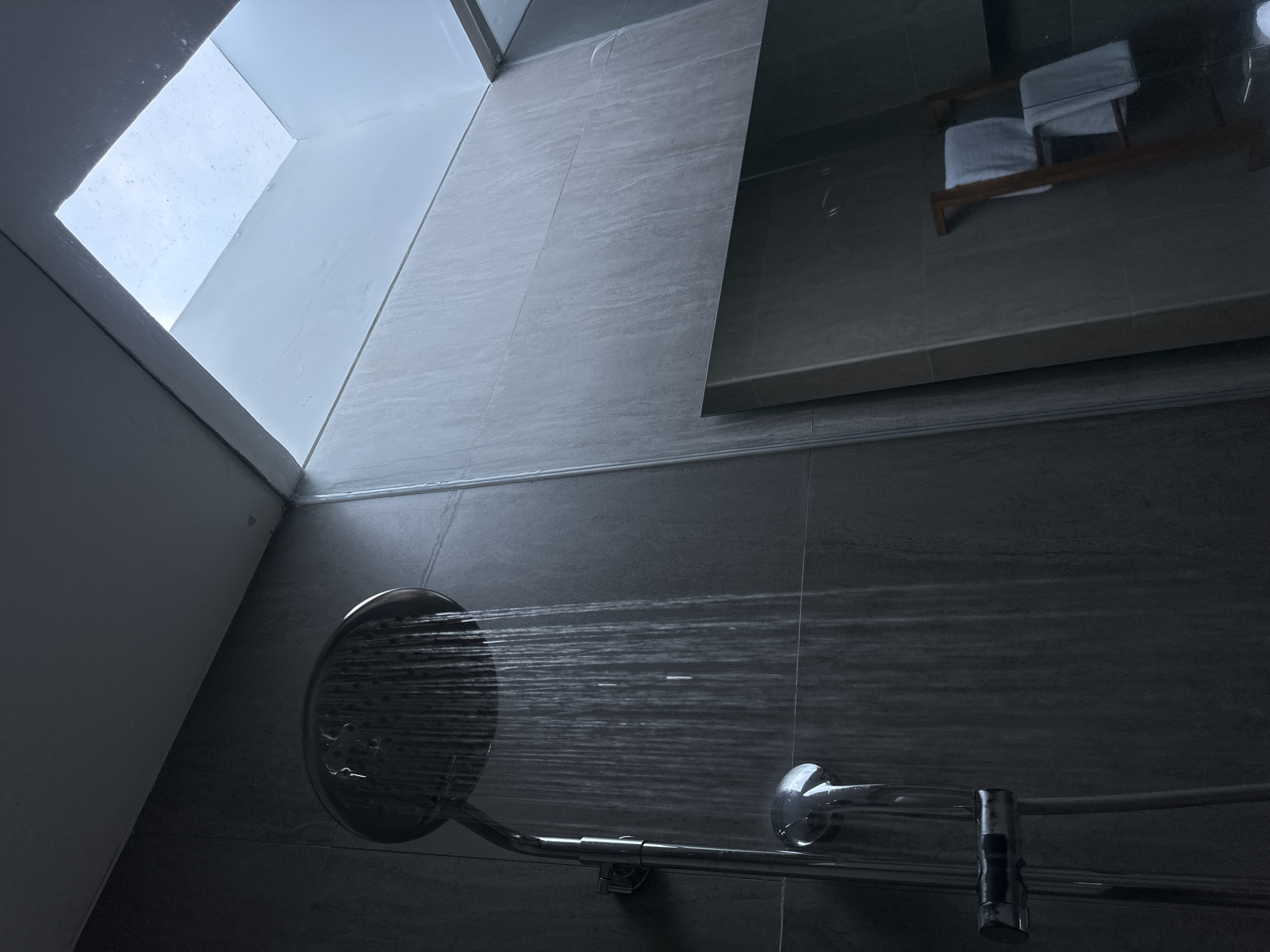 A low-angle, interior shot depicts a shower in a bathroom. The showerhead is mounted on a horizontal pipe with water streaming from it. The bathroom walls are tiled with gray, horizontal, rectangular tiles that mimic a wood grain. The upper left corner of the shot shows a skylight in the ceiling. The upper right shows a glass panel of a shower stall, with a blurry reflection of a towel rack with white towels. The scene takes place indoors, in a presumably private residence or hotel room, in an unknown location in Sukawati, Indonesia.