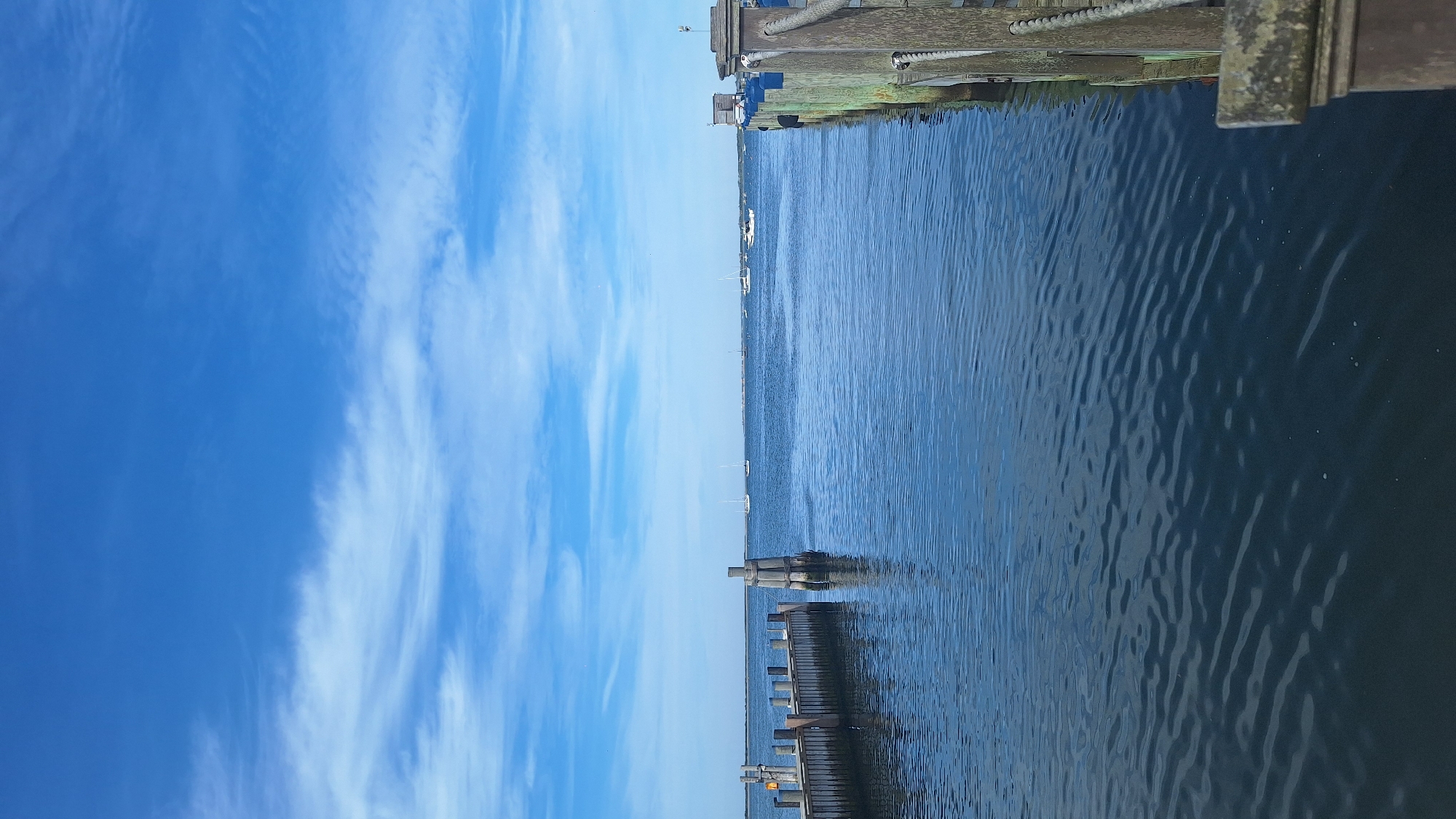 This is a daytime, eye-level, vertical, full shot of a body of water with a dock in the foreground and sailboats in the distance. The sky is bright blue with sparse white clouds. The water is calm with gentle ripples. On the left side of the image, a wooden dock extends into the water, with several bollards. In the center of the image, a thick wooden post rises from the water. On the right side of the image, another wooden dock is visible. Several sailboats are scattered across the horizon. The scene appears to be a harbor or bay. The location is Nantucket, United States of America.