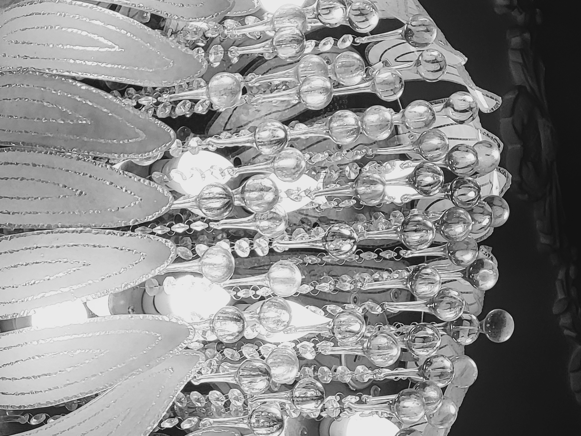 The image shows a close-up, black and white photograph of a chandelier. The chandelier consists of multiple rows of round, clear glass beads strung together, interspersed with leaf-shaped elements. Behind the beads are light bulbs, creating bright spots in the image. The background is dark and indistinct. The image does not provide enough information to determine the specific location in Yerevan, Armenia.