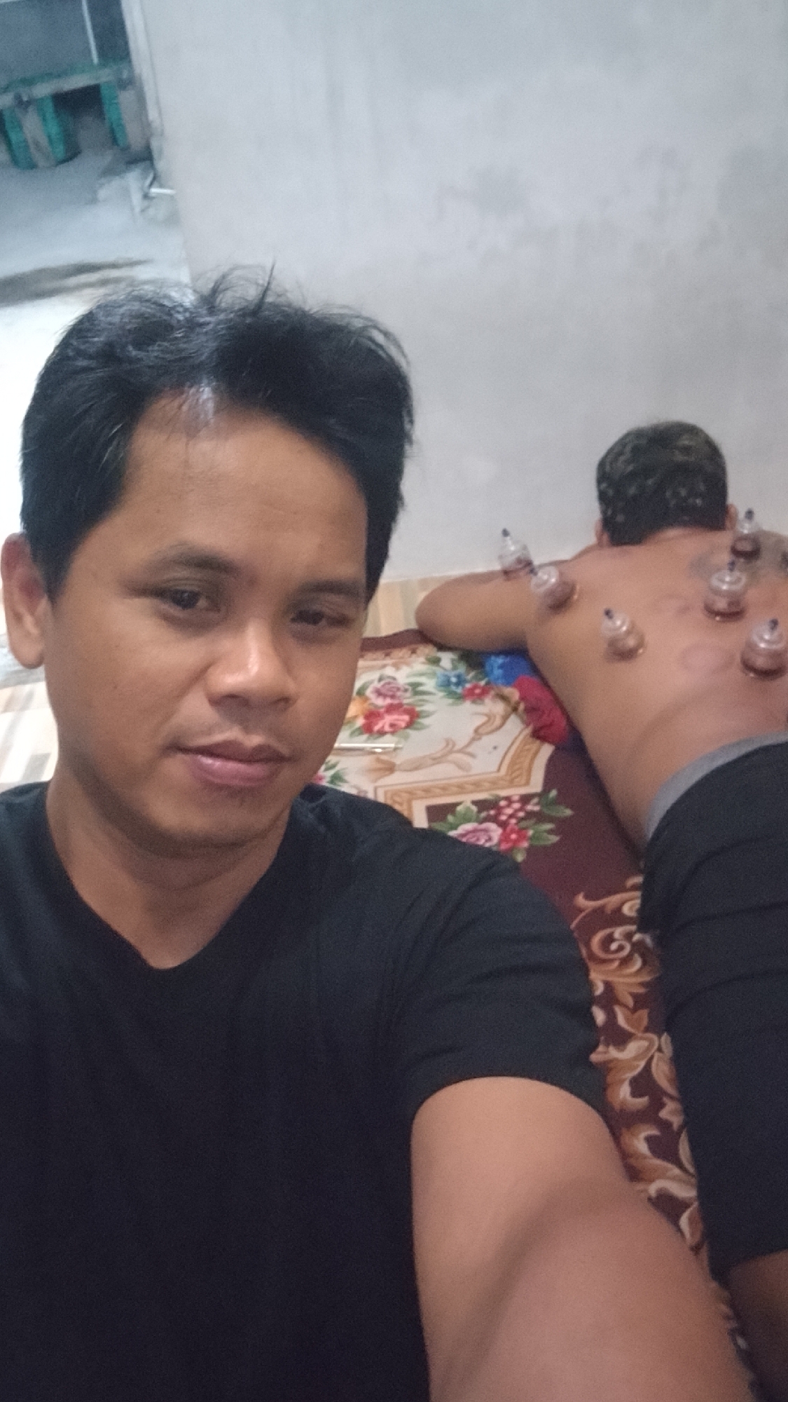 A man in a black t-shirt is taking a selfie in the foreground, with another person lying face down on a patterned mattress in the background. The person in the background has multiple glass cups placed on their back, suggesting a cupping therapy treatment. The environment appears to be an indoor space, possibly a bedroom or treatment room, with a light-colored wall and a glimpse of what might be outdoor elements through an opening to the left. The overall lighting is somewhat dim. There are no visible identifiers for the specific location within Karanglangu, Indonesia.