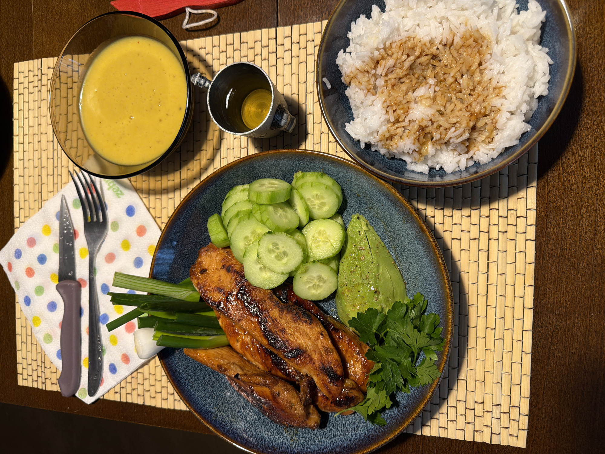 The image displays a meticulously prepared meal, set on a dark wooden table atop a light-colored woven placemat, likely in an indoor setting in Varna, Bulgaria.
The central dish is served on a dark blue/teal plate, featuring several pieces of what appears to be grilled and glazed chicken or fish, identifiable by its shiny, caramelized surface and distinct char marks. This main protein is accompanied by neatly sliced cucumbers, half an avocado, a vibrant bunch of fresh parsley, and several stalks of green onions.
To the upper right, a matching dark blue/teal bowl holds a portion of white rice, generously drizzled with a dark brown, savory sauce or dressing. On the upper left, a clear glass bowl contains a creamy, yellowish sauce or dressing, possibly mustard-based, with visible specks of spices. Beside it, a small, ornate metallic cup holds a golden liquid, suggesting a drink like tea or an aperitif.
Utensils, a knife with a grey handle and a silver fork, rest on a white paper napkin adorned with multicolored polka dots (red, blue, yellow, green). Partial text, "Zeb," is visible on the napkin. The lighting casts subtle shadows on the placemat, highlighting the texture of the woven material and adding depth to the overall presentation of the meal.