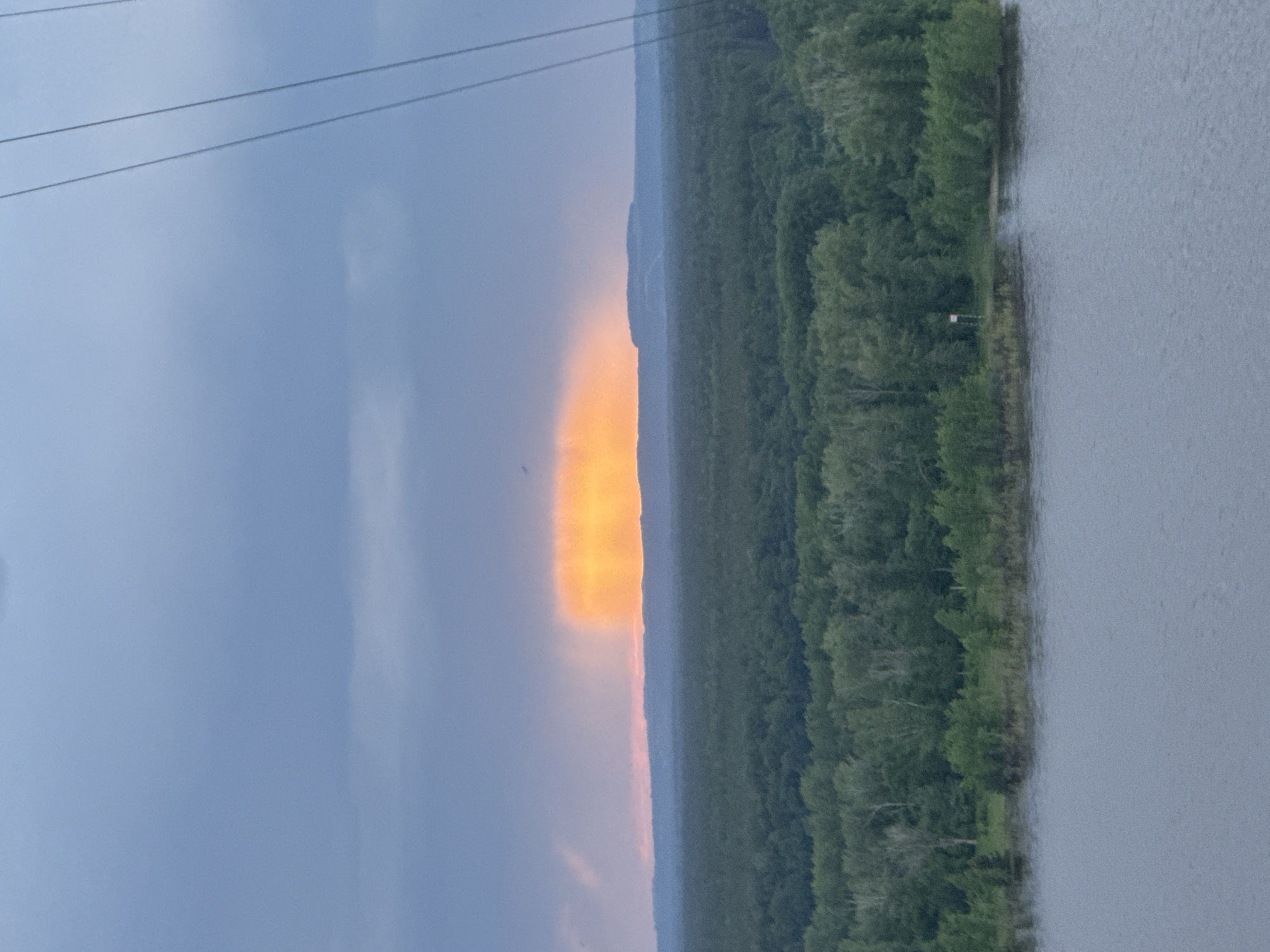 The image presents a landscape with a body of water, possibly a river or lake, on the right. A dense green forest lines the edge of the water. To the left of the forest, the sky transitions from a bright orange-yellow color near the horizon to a gray-blue hue higher up. A thin, dark silhouette of land is visible between the bright sky and the forest. Overhead power lines are visible in the upper left corner of the image. The city of Chaikovsky, Russia, is not readily apparent in the image, and there is no evidence of people or specific identifiable objects.