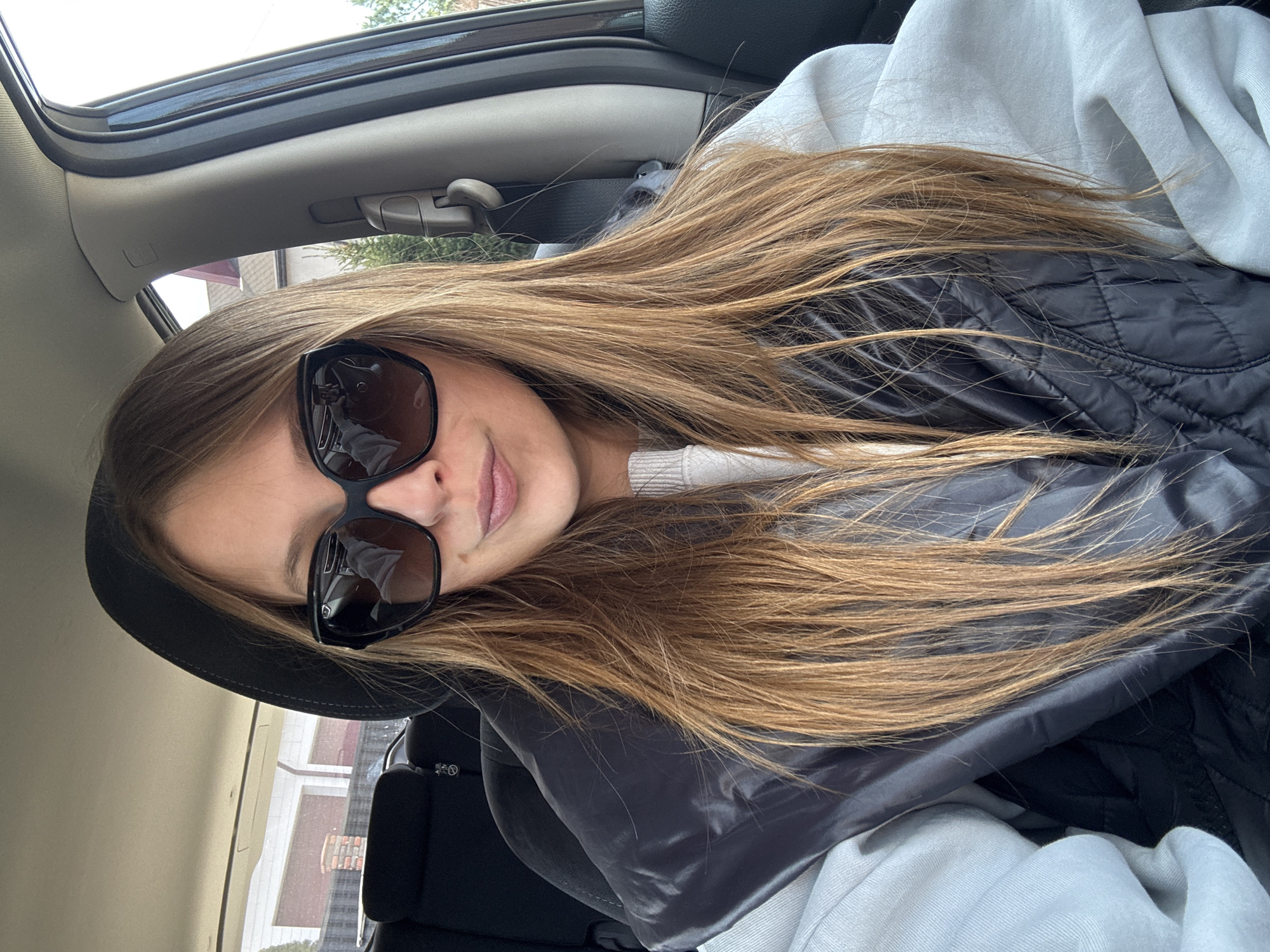 A young female sits in the passenger seat of a car. She has long, light-brown hair. She wears large, black sunglasses. She wears a white sweater and a black quilted jacket. The interior of the car is visible, including the sunroof and side window. Outside the window, the exteriors of buildings can be seen. The location is in the city of Ждановіцкі сельскі Савет, Belarus.