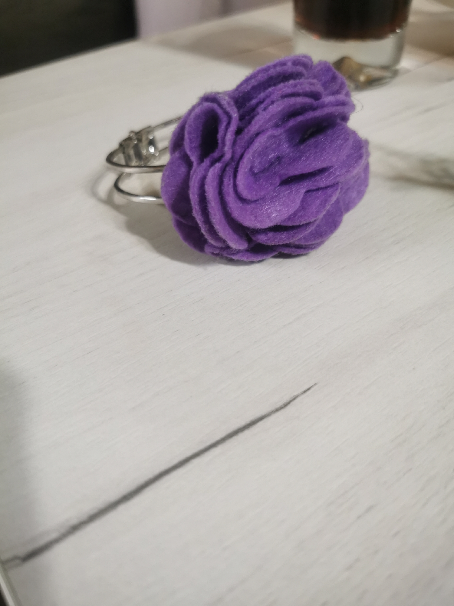A purple felt flower hair tie rests on a white, wood-grain surface. In the background, a partially visible glass containing a dark liquid sits on the surface. The location is unknown, but the city is Stara Zagora, Bulgaria.