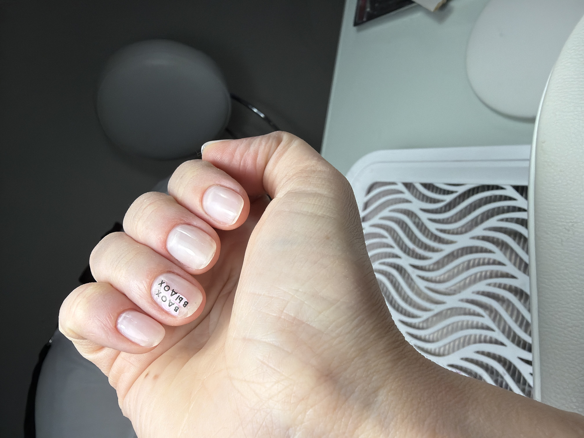 A close-up shot depicts a Caucasian woman's hand, positioned with fingers slightly curled, revealing neatly manicured fingernails painted with a light pink polish. Black lettering "BAOX" is mirrored and stacked on the rightmost fingernail. The hand rests against a light gray background, beside a white object with a wavy, cutout pattern. A rounded lamp is faintly visible in the background. The location cannot be determined from the information in the image.