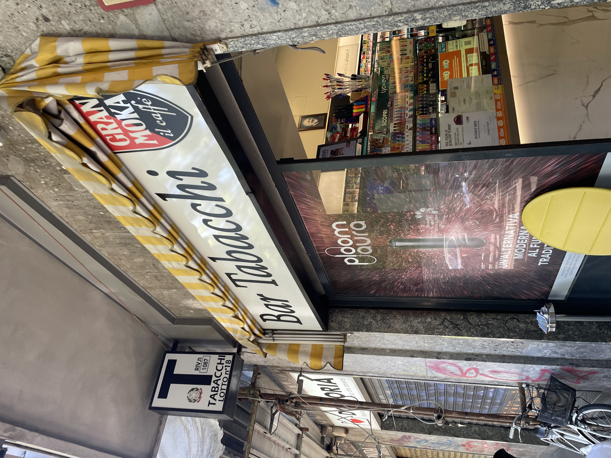 The image shows the exterior of a Bar Tabacchi (bar and tobacco shop) in Milan, Italy. The sign “Bar Tabacchi” is prominently displayed on a white awning with yellow and white stripes. The awning is labeled "Gran Moka il Caffè." Below is a smaller sign with a "T" symbol indicating “Tabacchi Lotto n°18.” Above the awning, is a poster advertisement for “ploom laura,” a smokeless heated technology. Inside the shop, various products including tobacco items, pens, and other miscellaneous goods are visible on shelves. A picture of an older man sits above the items for sale. The sidewalk is visible below, along with a bicycle. Other shops are also visible in the background.
