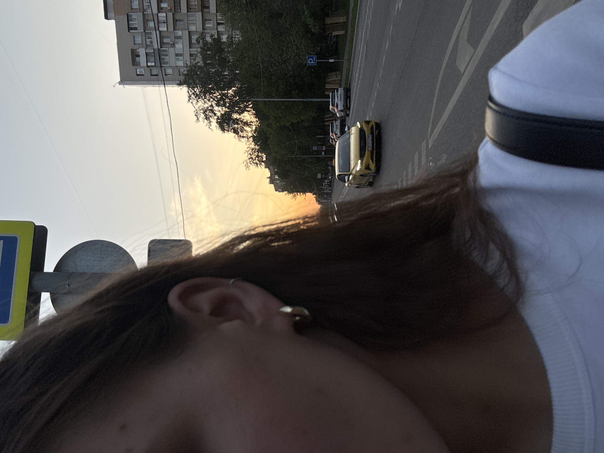 A photograph taken from a personal perspective shows a view along a street in Moscow, Russia, at sunset. A yellow car is on the road, and a tall apartment building is visible in the background. The photographer's ear and shoulder are in the foreground.