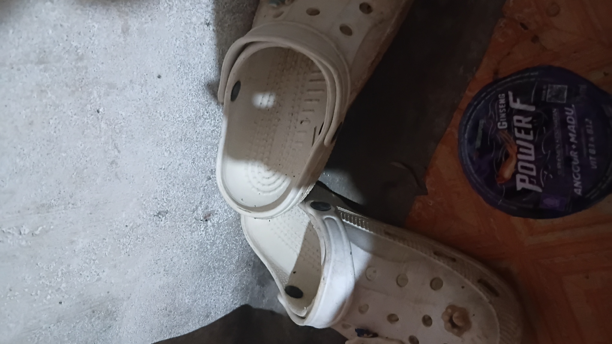 Two white Croc-style clogs are positioned on an orange tiled floor, with their soles facing upwards. One clog is in the foreground on the left, and the other is slightly behind and to the right, overlapping the first. Both clogs appear to be dirty and worn, with small dark specks visible on their surfaces. To the right of the clogs, a dark purple circular container is placed on the floor. The container has a label with text and an illustration. The text "POWER F" is prominently displayed in white letters, above smaller text that includes "GINSENG", "ANGGUR + MADU", and "VIT B3 + B12". The background consists of a textured, light-colored wall. This scene is located in the city of Prabumulih, Indonesia.