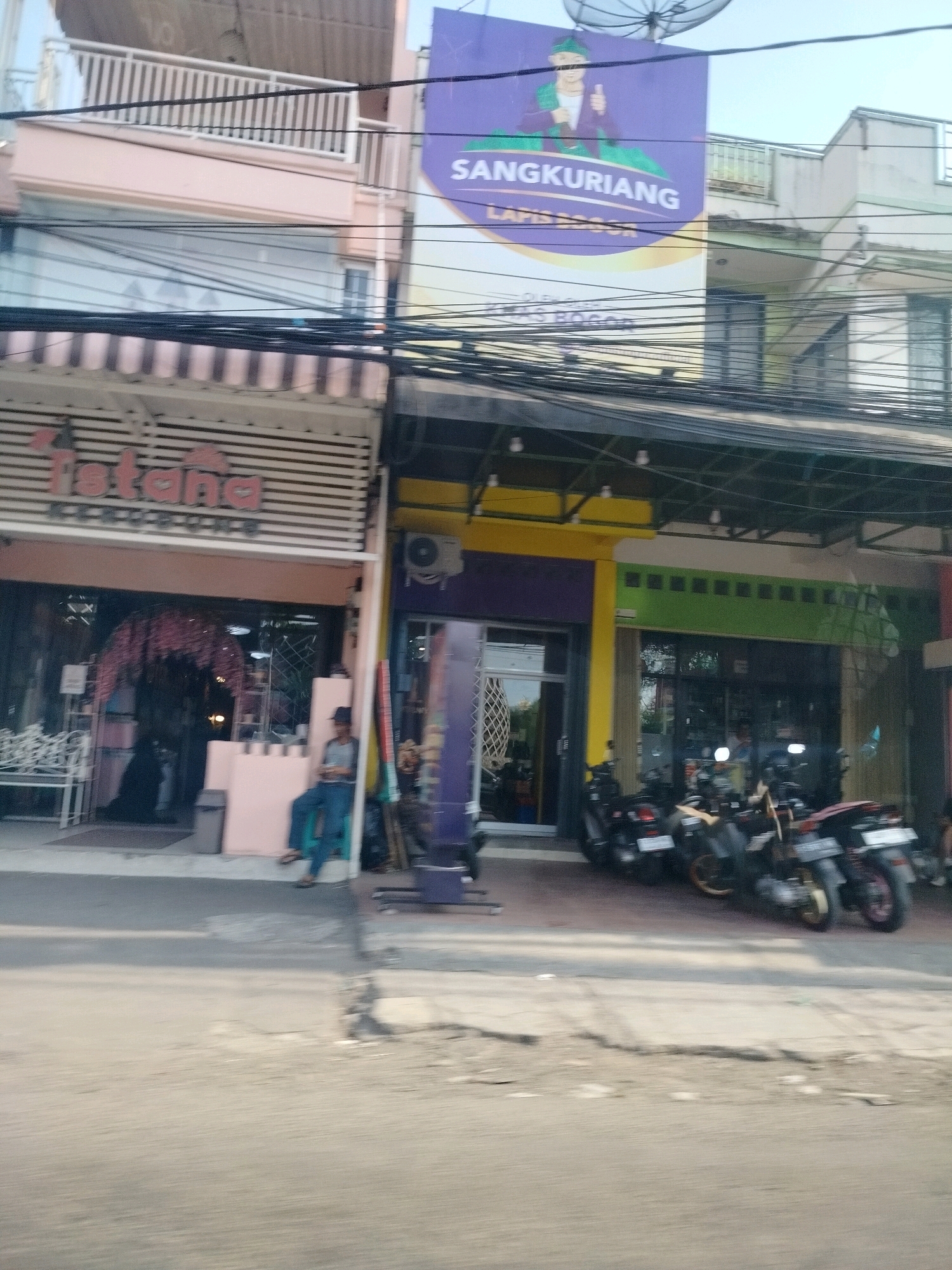 The image shows a street view in Cisarua, Indonesia, with several shops and buildings lining the road. In the foreground, a street with asphalt is visible, with some debris and cracks. On the right side of the street, a row of motorcycles are parked.
The buildings in the background house various businesses. A prominent sign with purple and yellow colors advertises "SANGKURIANG" and "LAPIS BOCOR". Next to it, a shop with a pink facade has the name "Istana" in red lettering above its entrance. The entrance features a dark interior with displays and a seating area. A person in blue pants and a dark shirt is seated on a low wall outside this shop, holding a dark object.
Further down the street, other shopfronts are visible with different colored facades, including yellow and green. These shops also have windows and entrances, with some motorcycles parked in front of them. An air conditioning unit is mounted on the exterior wall of one of the buildings. The overall scene depicts a busy commercial area with elements of daily life.