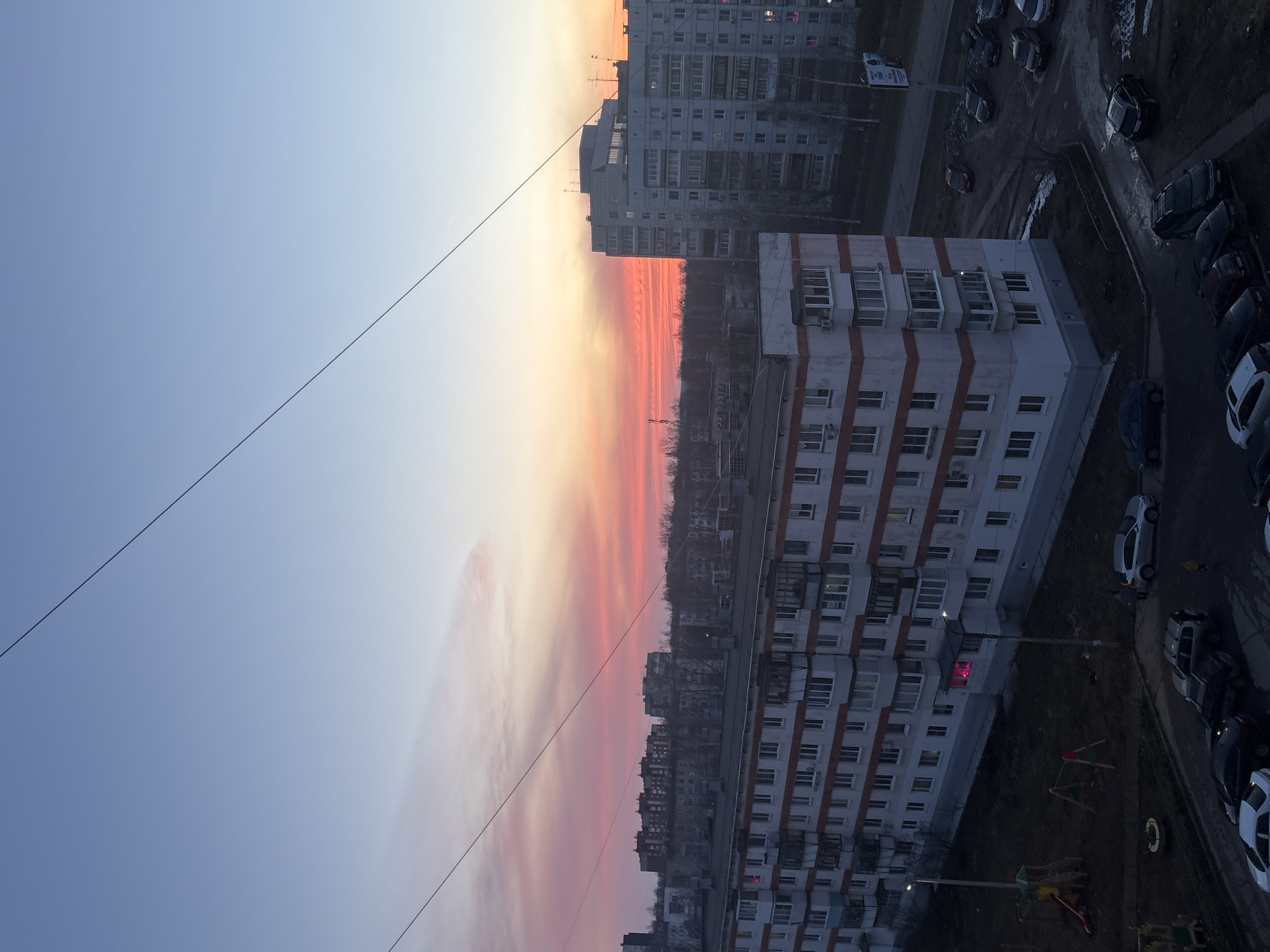 The image shows a cityscape at sunset in Chaikovsky, Russia. The sky is transitioning from a light blue on the left to a gradient of orange, red, and pink toward the horizon. Silhouetted buildings and trees are visible against the colorful sky. In the lower right foreground, there are multi-story white buildings with rows of windows, surrounded by parked cars and a small playground. A single power line crosses the sky diagonally from the upper left corner towards the center. Another multi-story building is visible in the upper center of the frame.