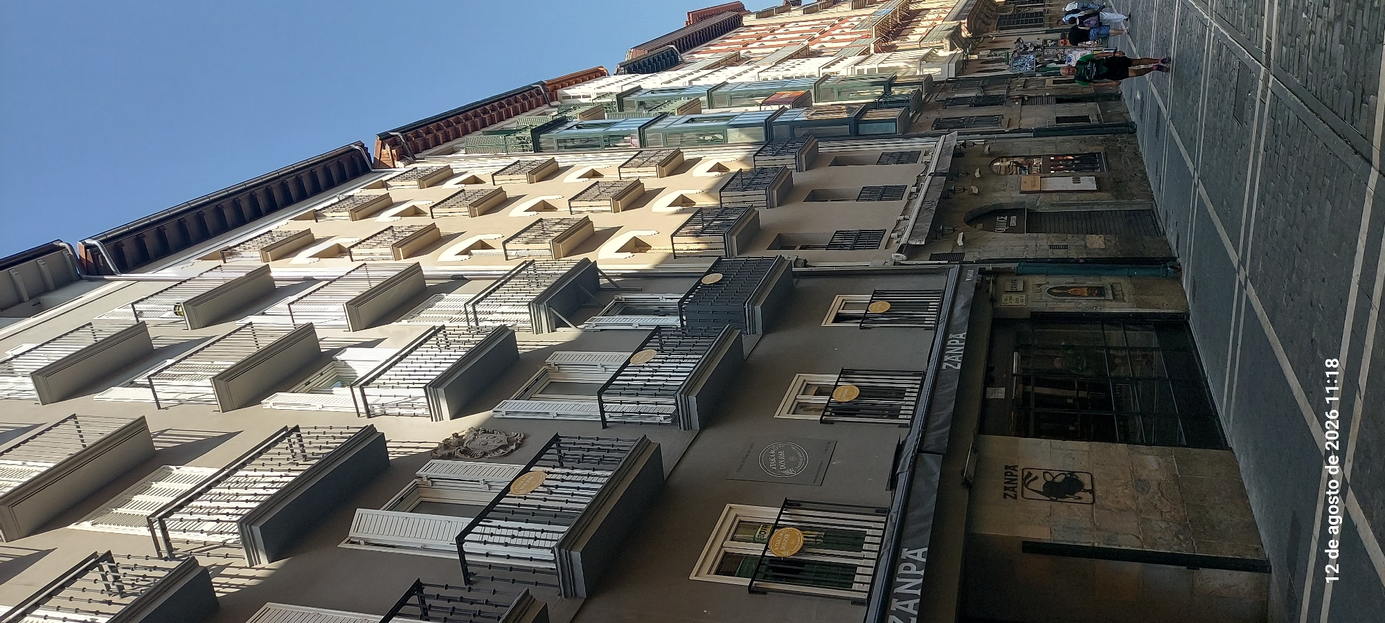 The image shows a street scene in Pamplona, Spain, on August 12, 2026, at 11:18 AM. The perspective is from ground level, looking up along a street lined with multi-story buildings. The buildings have a mix of architectural styles, with balconies, shuttered windows, and some modern bay windows. The street is paved with stone and marked with white lines.
On the left side of the street, a building facade is predominantly a dark grey color. It features several levels of balconies with black wrought-iron railings. On the ground floor, there is an awning with the name "ZANPA" printed in white letters. Below the awning, there is a stone facade with a large dark-colored entrance. A black metal silhouette of a bull is mounted on the stone facade.
Further down the street on the right, another building has a lighter beige facade with rows of balconies. Further still, there are buildings with colorful bay windows and a more traditional red-brick facade.
A few people are visible walking on the street. On the right side, a man in a green t-shirt and dark shorts is jogging. Behind him, a couple is walking away from the camera. The sky is clear and blue. The overall impression is a sunny day in a European city.