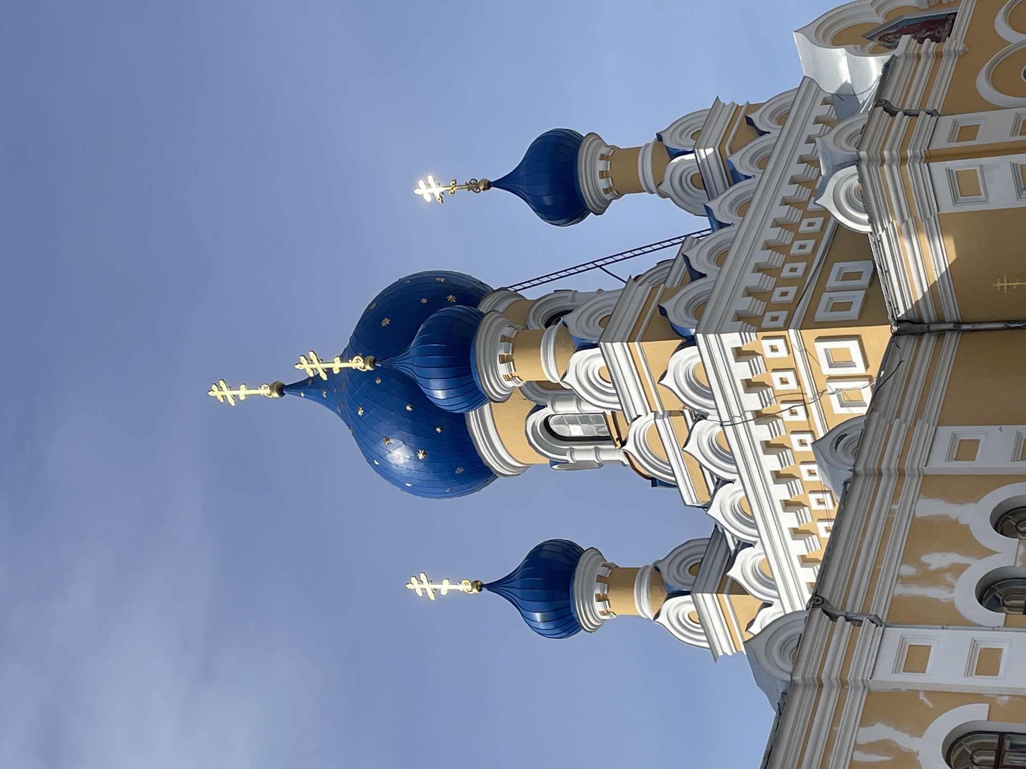 The image shows a close-up of the upper section of an Eastern Orthodox church, likely the St. Nicholas Garrison Cathedral in Brest, Belarus, due to its distinctive yellow and blue domes topped with golden crosses. The domes are painted a vibrant blue, some with decorative gold stars. The main structure of the church is painted a pale yellow with white decorative trim. Several arched windows are visible on the lower part of the building. The sky is clear and blue, with a few wispy clouds. There are no people visible in the image. The lighting suggests it is daytime, with sunlight glinting off the golden crosses.