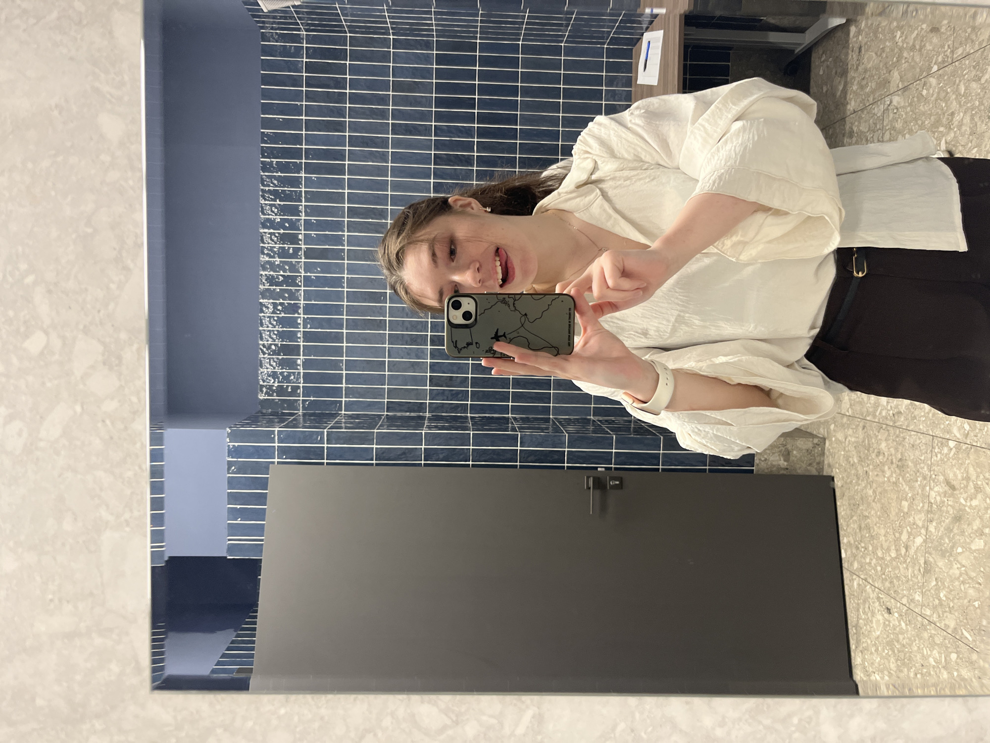 A young woman takes a mirror selfie in a bathroom with blue, horizontal tiled walls. The woman is wearing a white blouse and black pants. She holds a smartphone with a phone case depicting a map. The bathroom has a gray stall door with a simple latch. The floor is a light-colored speckled tile. The presence of tiled walls and a stall door suggests a public bathroom or restroom. The city of Moscow, Russia cannot be determined from the image.