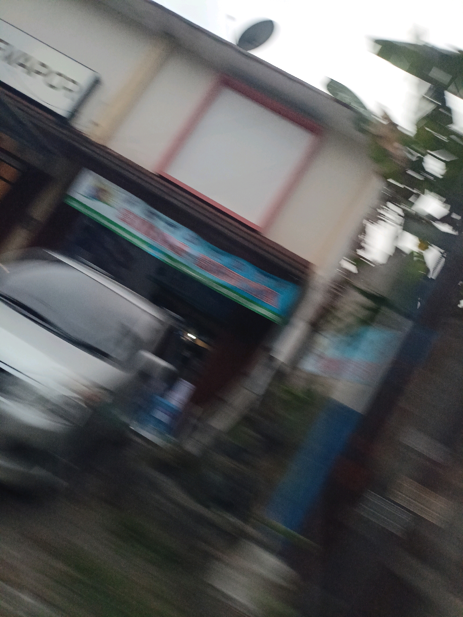 The image captures a blurred street scene in Magelang, Indonesia, likely taken from a moving vehicle. In the foreground, a white car is partially visible, its details obscured by motion blur. Behind the car, a building with a white facade and a reddish-brown trim is apparent. A sign with illegible text is mounted on the building, above a horizontally oriented banner with streaks of green, blue, and red. Further back, a blue vertical structure stands out, alongside foliage and other indistinct elements of the urban environment. The overall impression is one of a fleeting glimpse of a street in motion.