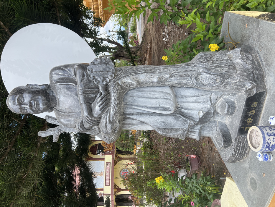 The image depicts an outdoor scene within a temple complex in Đăk Mar, Vietnam. A dark gray stone statue of a standing male figure occupies the central foreground. The figure is bald, has a beard, and exhibits a slight smile. It is depicted wearing long robes and holding a thick, gnarled tree trunk vertically against its chest with crossed arms. The statue is mounted on a dark gray stone base, which displays the inscription "TỔ HIỆP - TÔN - GIA". Directly in front of the base, a blue and white porcelain incense burner with a dragon motif, filled with ash, is visible, flanked by two smaller blue and white cylindrical containers.
In the background, to the left of the statue, a multi-story temple building with yellow walls, red-tiled roofs, and ornate golden and red decorative elements is partially visible. Surrounding the building are various green shrubs and potted plants, some bearing yellow and red flowers. A dense conifer tree canopy extends into the upper left quadrant. To the right of the statue, a large, white, circular disc is positioned behind its head. Further right, a large tree with a prominent trunk and exposed roots stands near a curving dirt path. A segment of an ornate golden structure is visible on the far right. The overall environment is green with diverse foliage under an overcast sky.