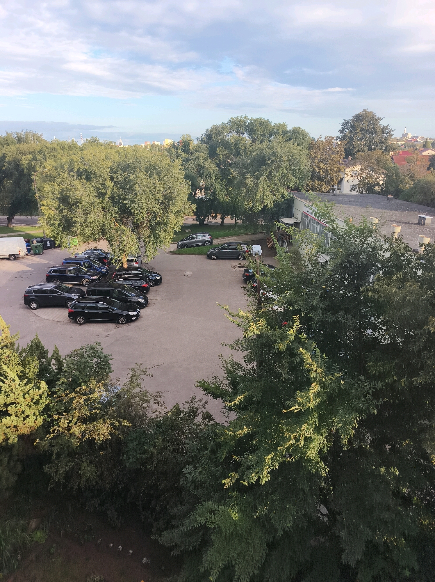 The image depicts an outdoor parking lot filled with several dark-colored cars, primarily SUVs and sedans. A white van is parked on the left side of the lot. The parking lot is surrounded by lush green trees and vegetation, with a building visible in the background on the right. The sky is partly cloudy with a mix of blue and white. The scene suggests a commercial or residential area with ample parking. The presence of trees and the architecture of the building indicate a European setting. The overall impression is one of a calm, everyday scene, possibly in a suburban or urban environment. The specific location is identified as Białystok, Poland.