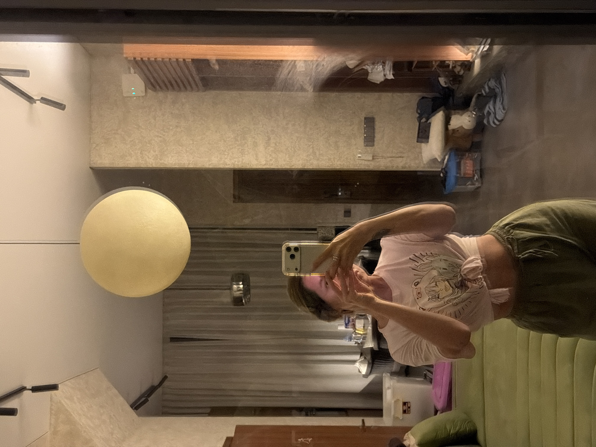A woman is taking a mirror selfie in what appears to be a modern, well-lit room. She is wearing a light pink t-shirt tied at the waist and olive green pants. The room features a large, round pendant light hanging from the ceiling, track lighting, and textured walls. In the background, a sofa, curtains, and various household items are visible. The reflection in the mirror shows the woman holding a smartphone and looking at herself. The overall impression is of a domestic interior setting.