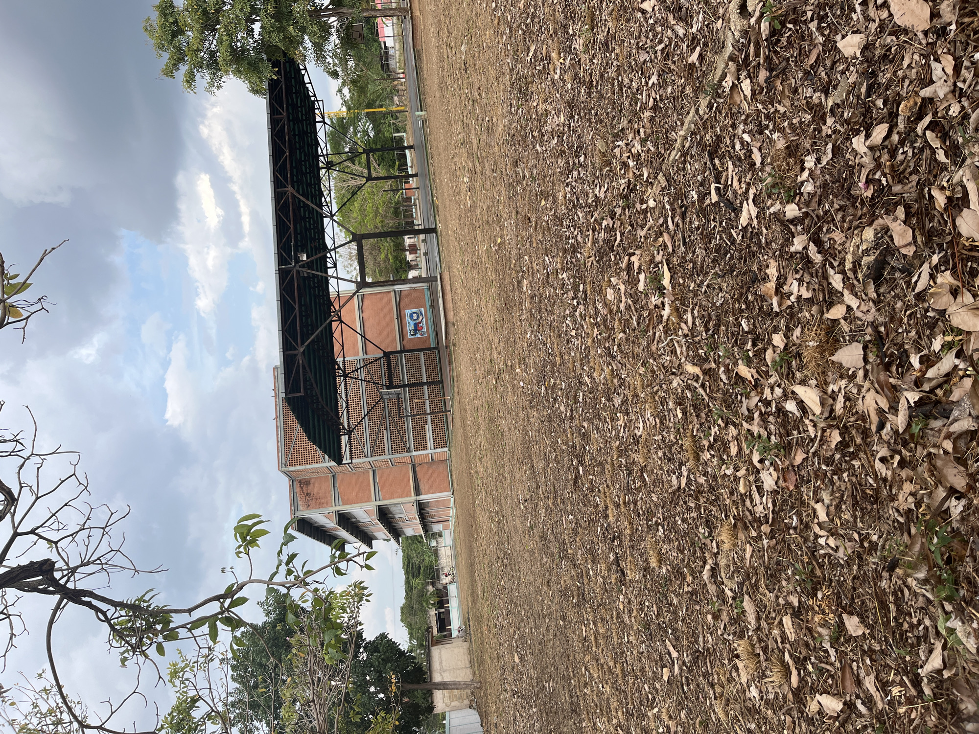 The image shows an exterior view in Tinaquillo, Venezuela. A large, red brick building with an attached metal structure dominates the scene, with a painted design on the side of the building. A field covered in dry leaves and vegetation extends to the right. In the foreground, branches of trees are visible. The sky is cloudy.