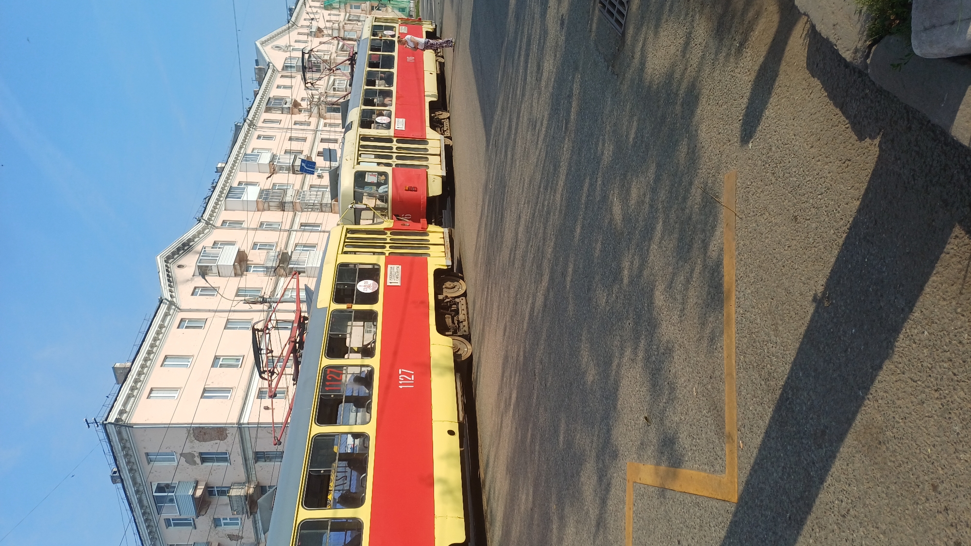 Two red and yellow trams are parked side-by-side on a street in Izhevsk, Russia, under a clear blue sky. The tram on the left is numbered 1127 and is closer to the viewer. The tram on the right is partially visible and appears to be numbered 76. Both trams have their pantographs raised, connecting to overhead power lines. In the background, a light pink, multi-story building with numerous windows and balconies lines the street. Shadows of trees are cast across the asphalt road. A person in white pants and a dark top stands near the rear of the tram on the right, facing away from the camera. Yellow lines mark a pedestrian crossing on the road. The scene depicts a typical urban streetscape with public transportation.
