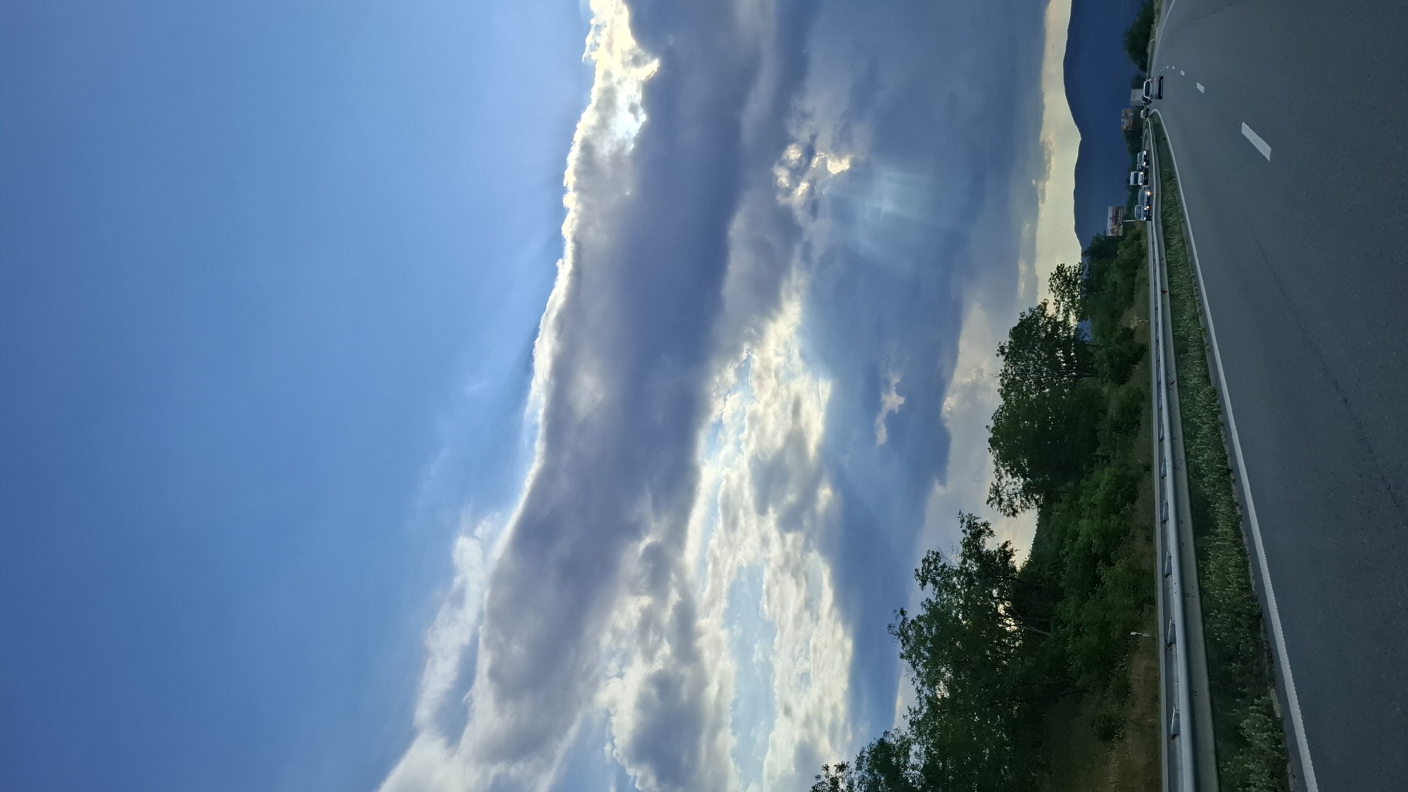 The image shows a view from a highway with vehicles on it. The highway has multiple lanes and a metal guardrail separating it from the side of the road. On the left side of the highway, there are green trees and vegetation covering a hillside. The sky is blue with dramatic clouds, and sunbeams are visible breaking through the clouds. In the distance, there are mountains. The scene appears to be a scenic view along a road in the city of Gorno Varshilo, Bulgaria.