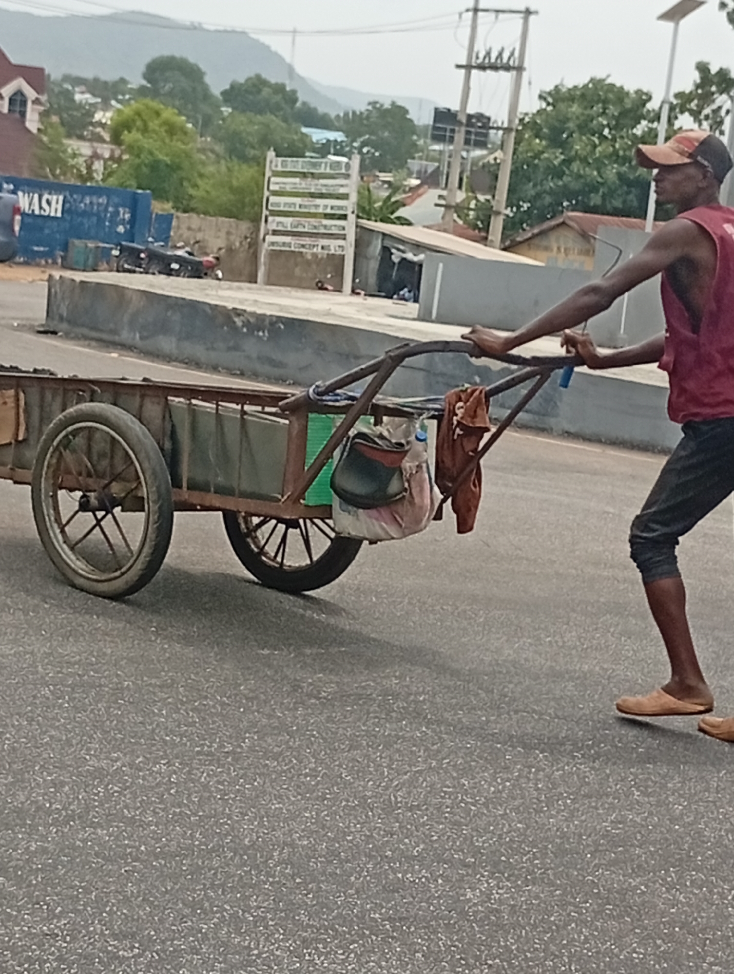 A man is depicted pushing a two-wheeled metal handcart along a paved road in Adankolo, Nigeria. He is wearing a red sleeveless top, dark cropped trousers, light-colored flat shoes or sandals, and an orange baseball cap. The handcart appears well-used, featuring mismatched wheels, and carries items including a green container, a multi-colored bag, a brown cloth or towel, and a plastic bottle.
The scene unfolds during daylight hours, under what appears to be an overcast or hazy sky, with mountains and lush greenery visible in the distance. In the background, there is a mix of urban and natural elements, including a building with a red roof and a prominent blue industrial container labeled "WASH." Power lines and poles are also visible. A white signboard in the mid-ground, though partially obscured and somewhat blurry, displays text including "OGI STATE GOVERNOR'S OFFICE," "RIVER STATE MINISTRY OF WORKS," "PALL GARY CONSTRUCTION," and "INSURGE CONCEPT NIG. LTD." The man's posture suggests he is actively engaged in moving the handcart, focused on his task.