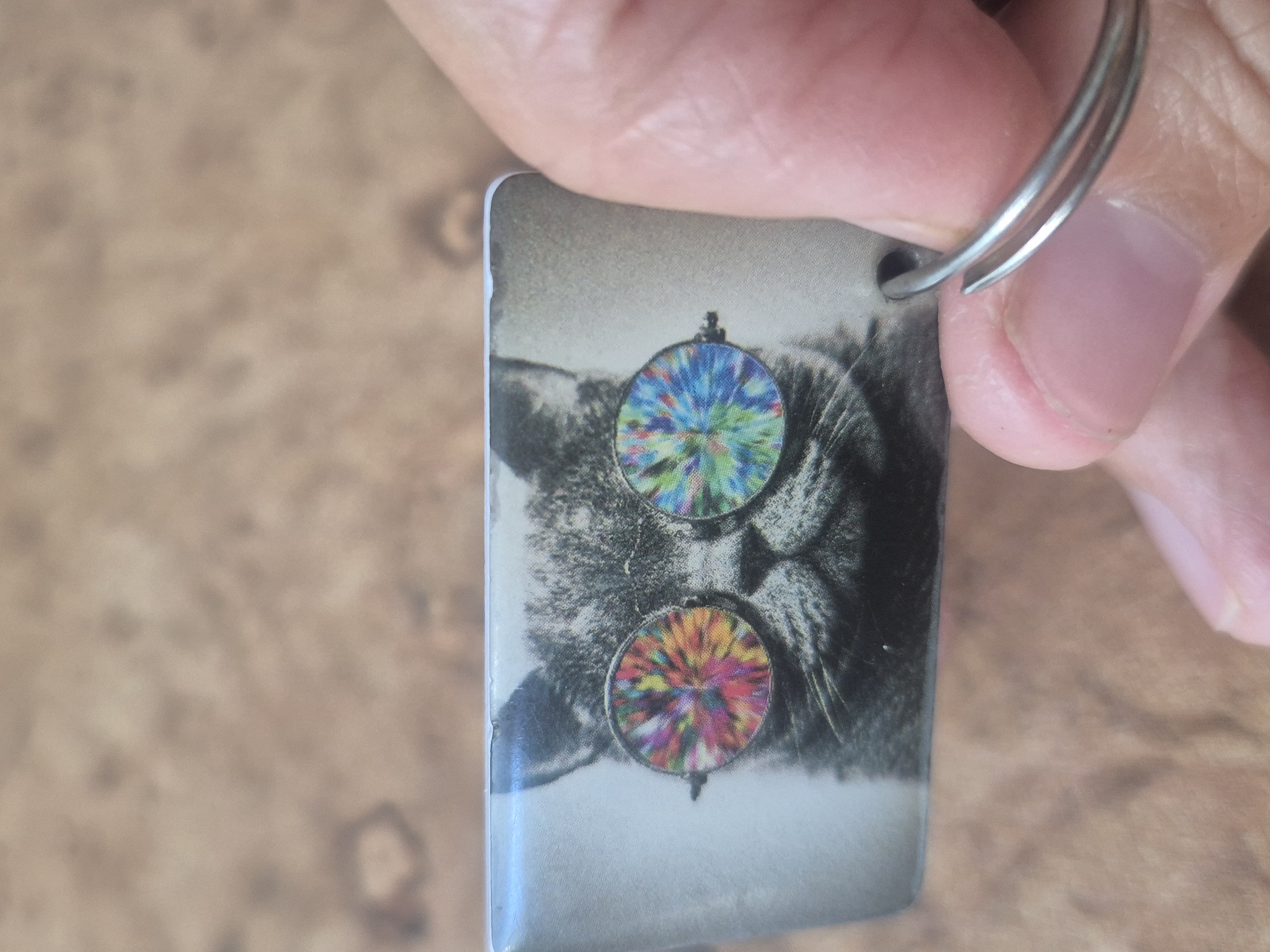 The image shows a close-up of a keychain featuring a grey cat wearing tie-dye sunglasses. The keychain is held by a person's hand, with the thumb and index finger visible. The background is a blurry, textured surface that appears to be wood. The image does not contain any information about the location of Kokshetau, Kazakhstan.