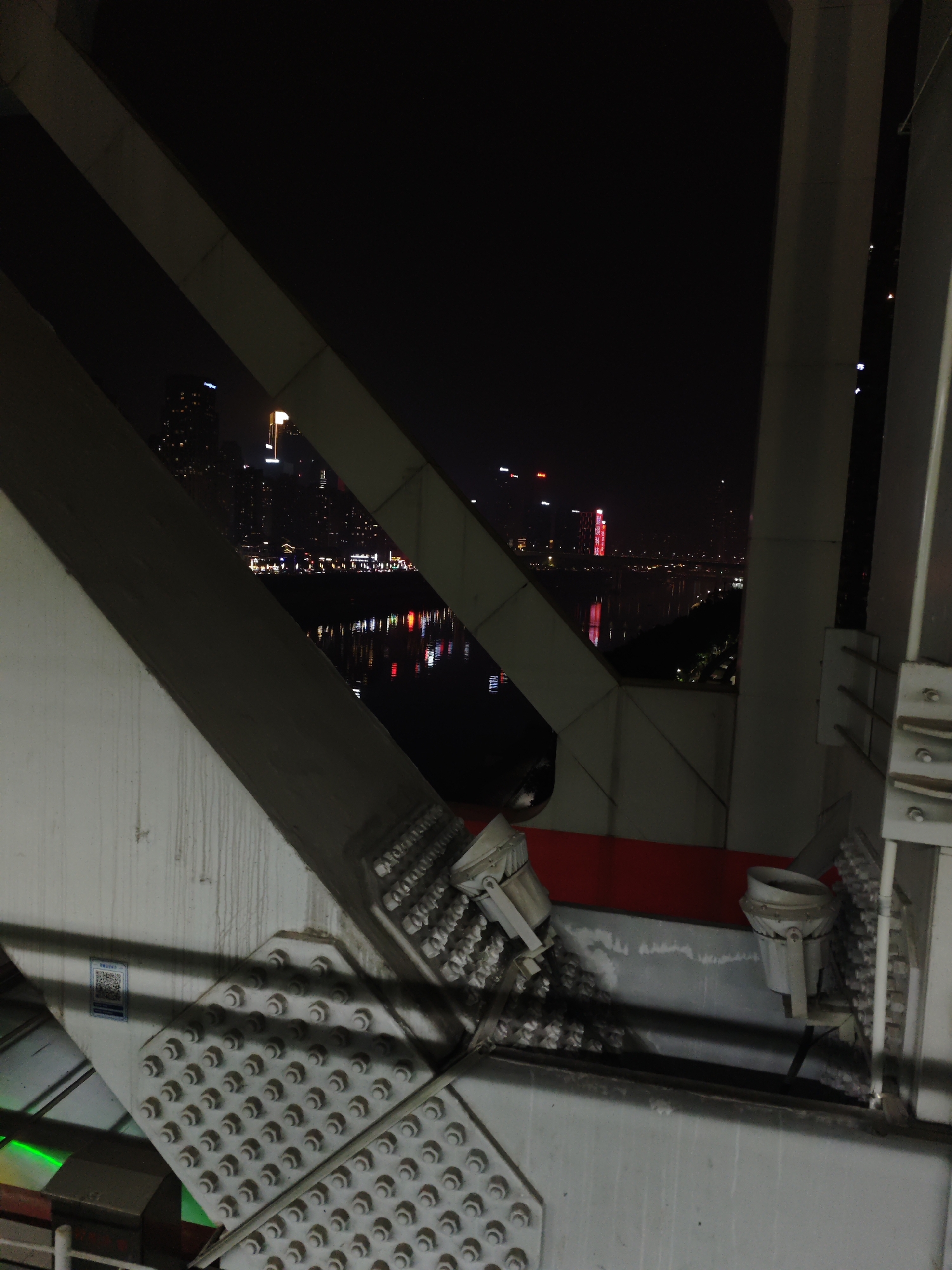 View from a bridge structure in Yuzhong District, China, at night. The structural beams are gray and are connected by bolts. Behind the beams, a cityscape is visible with lighted buildings and reflections on a river.
