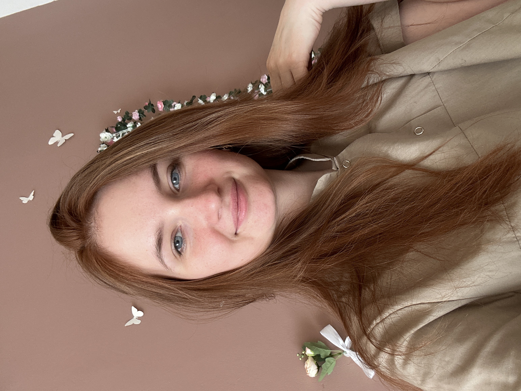 A young woman with fair skin and light red hair is centrally framed in a portrait-style shot. She has blue eyes and is smiling subtly. The background is a solid, muted brown. Her hair is arranged around her face and shoulders, with a floral headband partially visible at the top edge of the frame. Two small white butterfly decorations and a miniature floral bouquet tied with a white ribbon are situated in the lower left and right corners of the image respectively. She is wearing a light brown button-down shirt. The setting cannot be determined to be the city of Chaikovsky, Russia from the image.