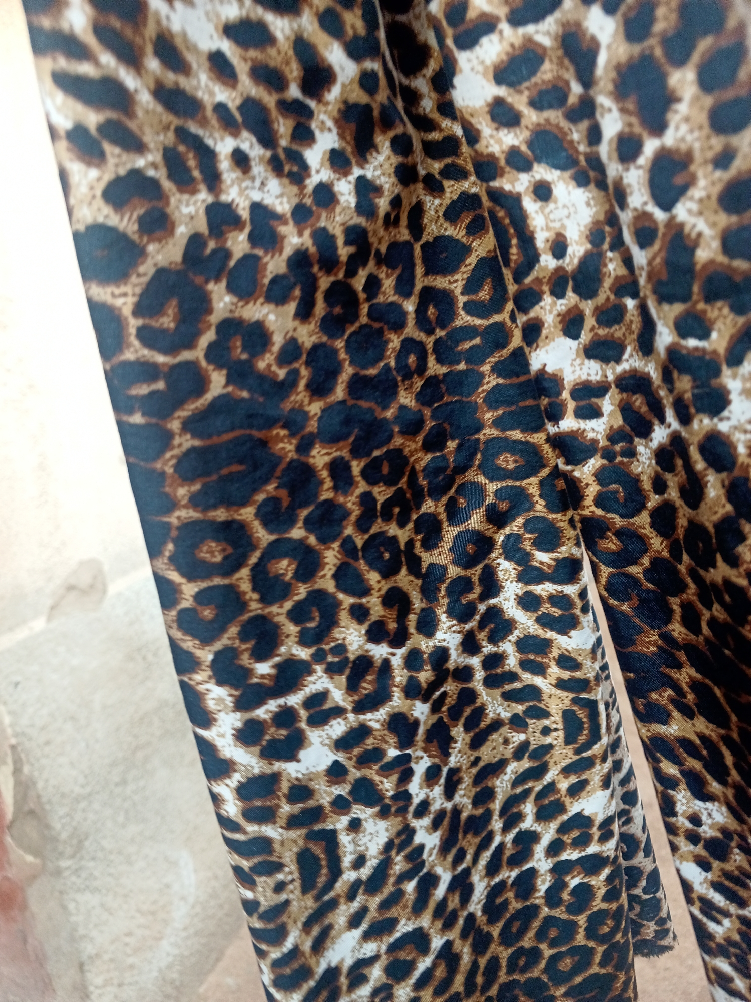 The image shows a close-up of a leopard print fabric, draped and folded. The fabric has the characteristic irregular spots of black and brown on a lighter tan or white background. The texture appears to be smooth and possibly synthetic, with some sheen. The fabric is illuminated from the side, casting subtle shadows that highlight the folds and the texture of the print. The background to the left of the fabric appears to be a rough, textured wall, possibly made of concrete or stone, with some discoloration and marks. There are no people, animals, or discernible landmarks present in the image that would allow for specific identification of a location in Jalingo, Nigeria. The image is a still life focused entirely on the textile.