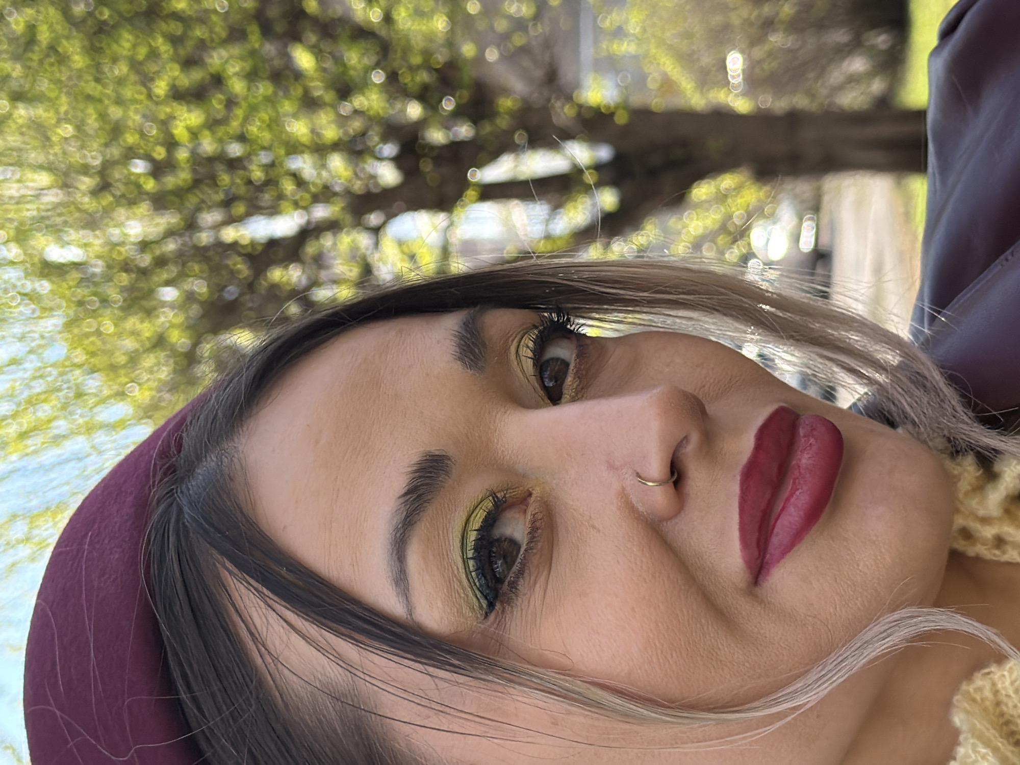 A close-up, portrait-style shot focuses on a woman's face. She is looking slightly upwards and to her right. The woman has fair skin, dark hair, and wears a maroon-colored headband. Her makeup is noticeable with yellow eyeshadow and dark red lipstick. She also sports a gold nose ring.
The background is blurred, featuring trees with green leaves. It is not possible to verify whether the location is Krasnoyarsk, Russia, from the image.
