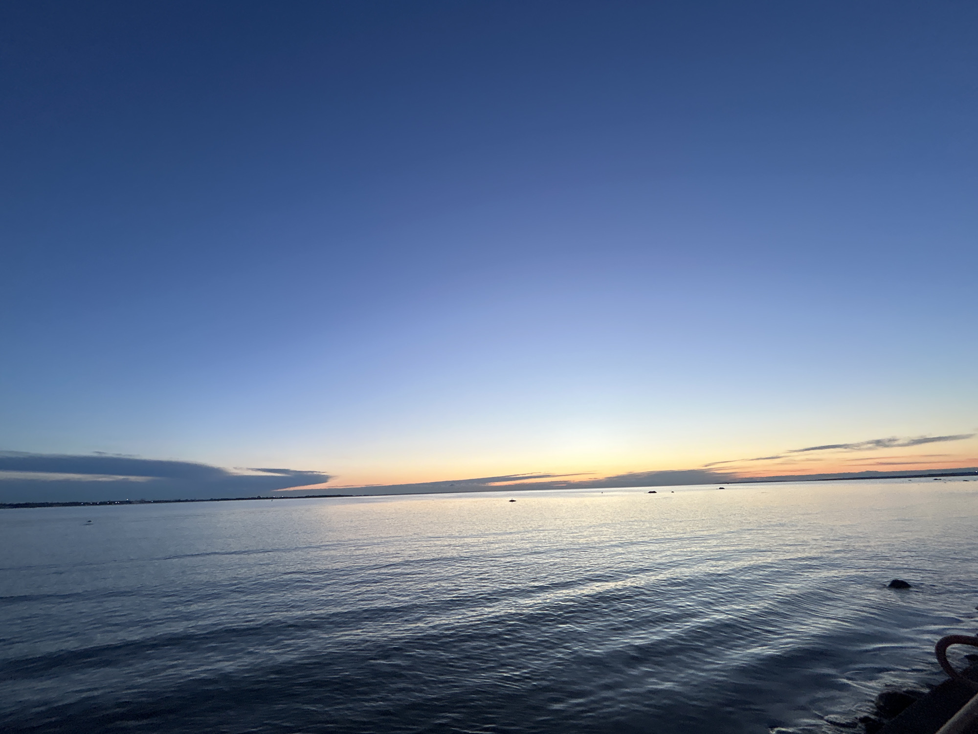 The image shows a body of water with ripples under a clear blue sky transitioning to a warm orange hue near the horizon. The horizon line is marked by a distant landmass and a few small, dark objects in the water, possibly rocks or buoys. The overall scene depicts a serene seascape, likely at dawn or dusk, with no people or prominent man-made structures visible in the main frame. The composition suggests a natural landscape with emphasis on the sky and water. Given the context of Tallinn, Estonia, this could be a view of the Baltic Sea from a coastal area.