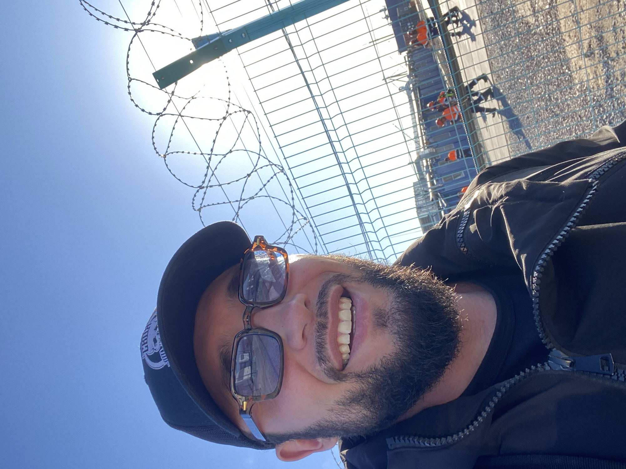 A man wearing a black baseball cap, patterned glasses, and a black jacket takes a selfie against a bright blue sky. He has a beard and is smiling. Behind him, a green metal fence with coiled barbed wire runs horizontally across the frame. Beyond the fence, several construction workers in orange safety vests and hard hats are visible, along with temporary buildings and shipping containers, suggesting a construction site. The overall scene appears to be outdoors during the daytime. The location is identified as the city of Koskolovo, Russia.