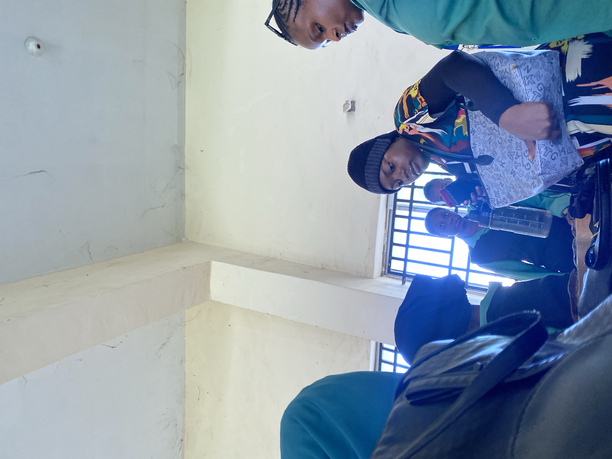 In an indoor setting, likely a classroom or common area in Rafin Par, Nigeria, a group of individuals are gathered. The environment appears somewhat weathered, with visible cracks on the ceiling and walls. Natural light filters in from windows with metal bars, suggesting a secure building. In the foreground, a young woman wearing a black beanie and a vibrantly patterned top is holding a water bottle and a patterned tote bag. She appears to be looking at something in the bag. To her right, another woman, possibly an adult, is wearing a green shirt and has her hair styled in braids. She is looking down and slightly away from the camera. In the background, other individuals are visible, one wearing a green shirt and another with a dark head covering. The overall impression is of a casual gathering or perhaps a break in an educational or community activity.