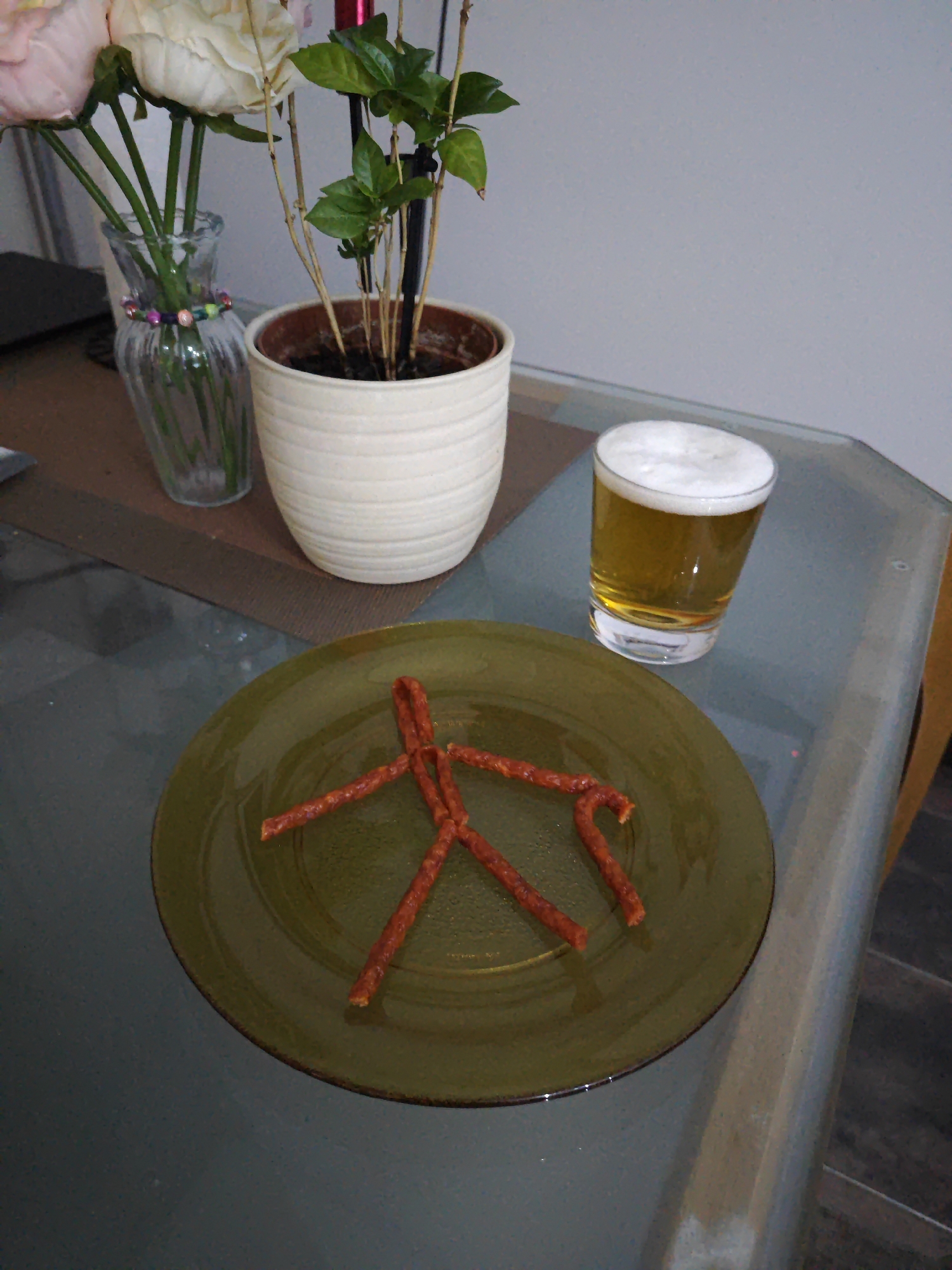 A still life image depicts a table setting with a plate of food and a drink. In the foreground, a green plate holds several pieces of thin, reddish-brown sausages arranged in the shape of a stick figure. To the right of the plate, a short, clear glass contains a pale yellow beverage topped with a frothy white head, likely beer.
In the background, a white ceramic pot holds a small plant with green leaves and bare branches. To the left of the pot, a clear glass vase with a decorative beaded band contains a bouquet of flowers, including a light pink one and a cream-colored one. The flowers appear to be roses.
The scene is set on a glass-topped table, with a dark brown placemat visible beneath the vase and pot. The lighting suggests it is daytime, possibly indoors given the lack of natural light cues. The overall impression is a casual, relaxed setting, perhaps a snack break or a moment of repose. There is no visible text in the image.