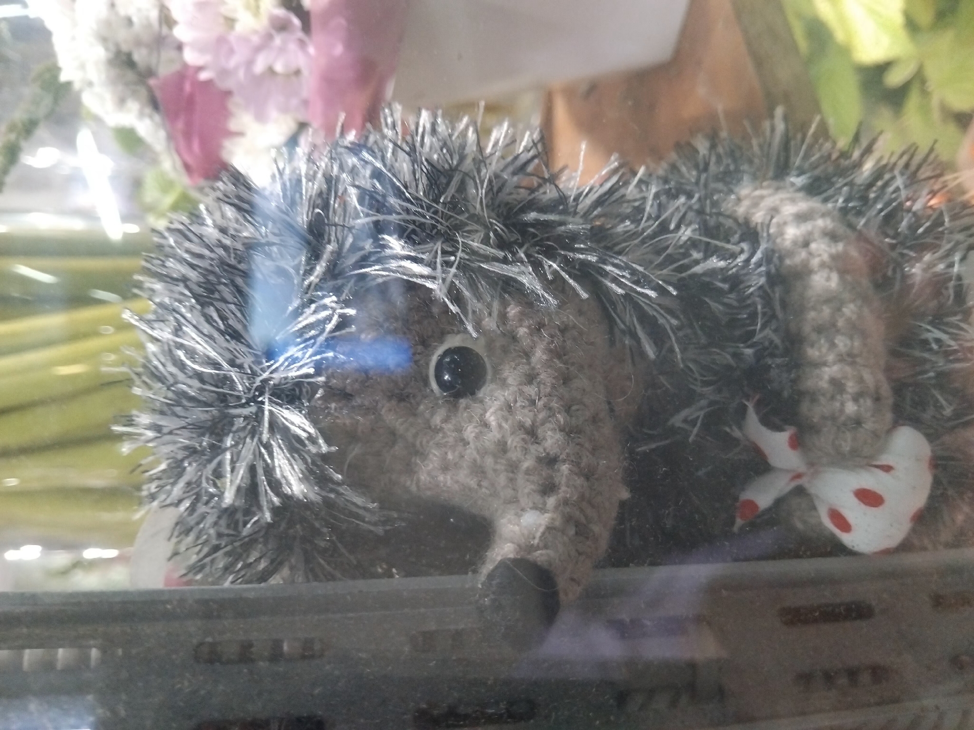 A close-up, eye-level, vertical, full shot shows a crocheted hedgehog figurine with black and silver yarn quills, positioned behind a clear pane of glass. The hedgehog is facing left, with its head tilted upwards and its black button eye visible. It is holding a small white bag with red polka dots in its paws. To the left of the hedgehog, a black, textured, vertical object with horizontal ridges is partially visible. In the background, out of focus, are some green, yellow, and pink floral elements. The lighting appears to be bright and reflects off the glass. The scene is consistent with a shop display.