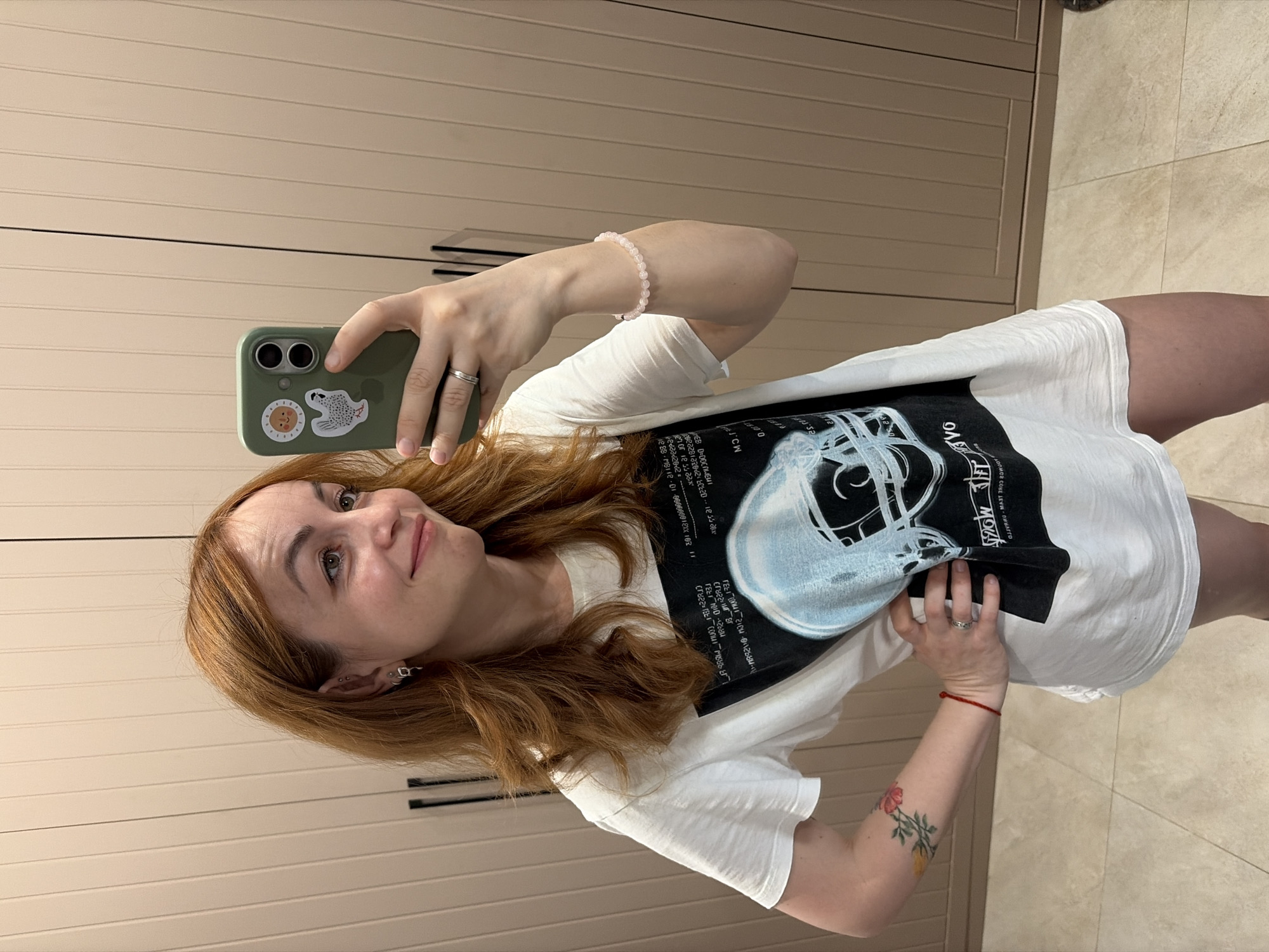 A woman takes a mirror selfie in an indoor setting, likely in Chaikovsky, Russia. She has reddish-brown hair and is looking towards her phone with a pleasant, slight smile. She is wearing a white short-sleeved t-shirt featuring a prominent dark graphic print on the front. The graphic includes a stylized white or light-colored outline of what appears to be a skull or helmet, surrounded by text and abstract designs, some of which are inverted.
Her left hand, adorned with a silver ring on the ring finger, holds an olive green smartphone with two visible camera lenses on the back. The phone case has two stickers: one circular with a smiling face, and another depicting an animal, possibly a seal or similar creature. On her right wrist, she wears a light pink beaded bracelet, and a red string bracelet is visible on her lower right forearm. A floral tattoo, appearing to be a rose with green leaves, is on her upper right arm. A silver-colored earring or piercing can be seen in her right earlobe.
The background consists of light beige or cream-colored paneled wardrobe doors with dark handles, suggesting a bedroom or dressing area. The floor is covered with light-colored tiles. Visible text from the t-shirt graphic, though partially inverted and stylized, includes what appears to be a serial number or product code like "1288-MB112-01-000000012XX102," and other fragmented or distorted words. The overall atmosphere is casual and relaxed.
