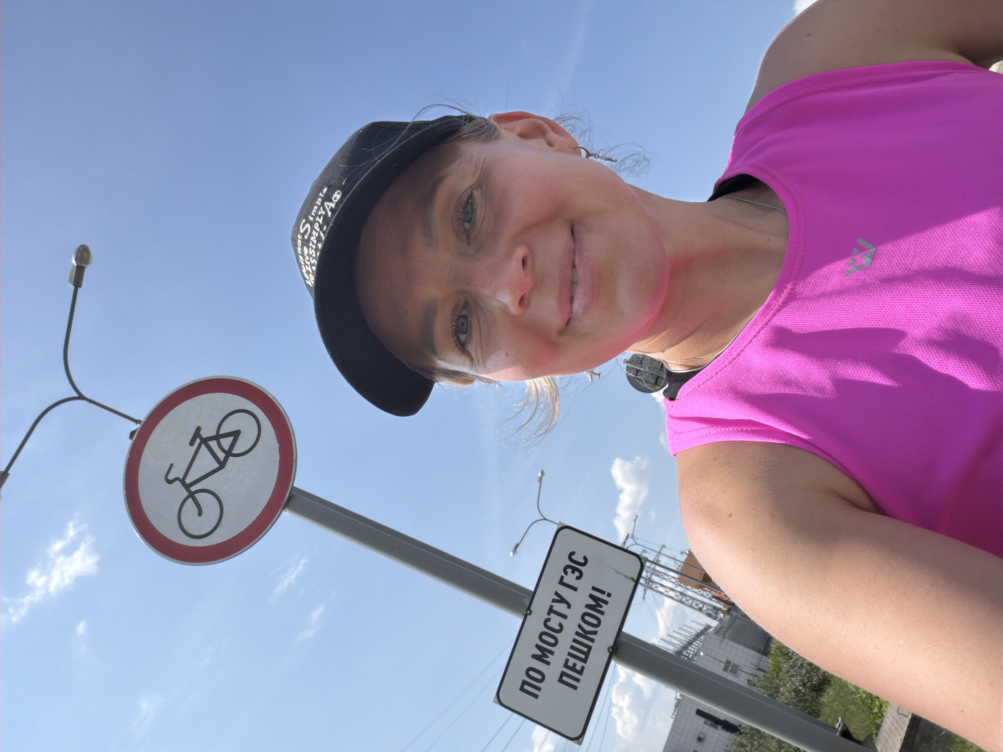 A woman is taking a selfie on a sunny day in Chaikovsky, Russia. She is wearing a bright pink athletic tank top and a black baseball cap. Behind her, a sign reads "ПО МОСТУ ГЭС ПЕШКОМ!", which translates to "On the HPP bridge on foot!". There is also a circular road sign depicting a bicycle crossed out, indicating no cycling is allowed. The background shows a blue sky with scattered clouds, power lines, and some industrial structures.