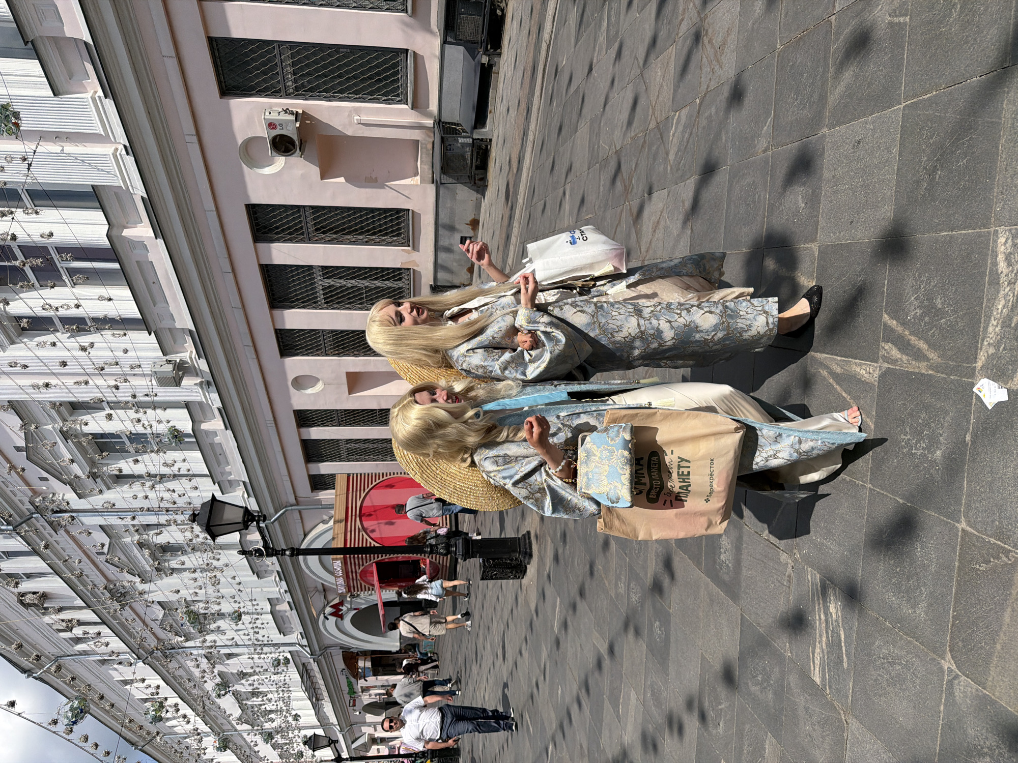 This outdoor street scene in Moscow, Russia, captures two women in the foreground, seemingly posing or engaging in a street event, under bright midday sunlight. Both women have long blonde hair (possibly wigs) and are dressed in striking, elaborate robes or kimonos featuring a light blue and gold/silver patterned fabric. One woman wears a wide-brimmed straw hat. Each holds shopping bags: the woman on the left has a brown paper bag and a patterned clutch, while the woman on the right carries a white bag.
The setting is a pedestrian street paved with dark grey square tiles. Buildings with multiple windows and grates line both sides of the street; one building features an LG air conditioning unit. Overhead, an extensive installation of strings adorned with numerous small decorative elements and reflective spheres creates an ornate canopy. Long shadows stretching across the pavement confirm a sunny day.
In the mid-ground and background, other pedestrians are visible, walking or standing. A black bollard is present on the street. Visible text on the brown shopping bag reads "ВМЕСТО ПАКЕТА — СБЕРЕЖЕМ ПЛАНЕТУ" (Instead of a bag - save the planet) and "ПЕРЕКРЁСТОК" (Perekrestok). Part of a "CHANEL" logo is visible on the white bag. A red circular sign with an "M" symbol (likely a Metro station) and some Cyrillic text are discernible in the distance, firmly placing the scene in Moscow. The women's distinctive attire suggests a performance, photoshoot, or a fashion-forward outing.