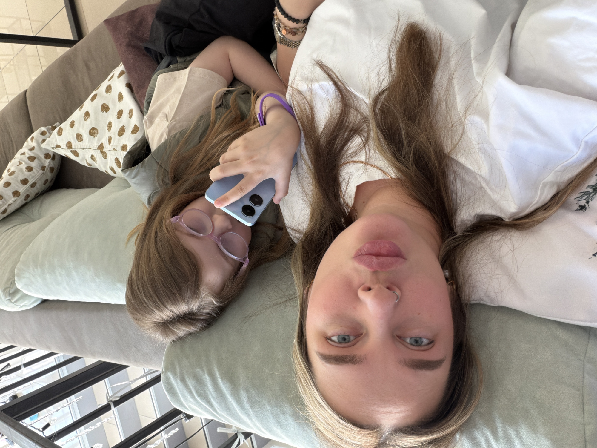 Two females are depicted in a close-up, upward-facing shot, likely a selfie, as they lie on a light-colored sofa or bed. The individual on the right, appearing to be an adult, has long light brown hair, blue eyes, and a septum piercing. She wears a white top and looks directly at the camera with a neutral expression.
Beside her, on the left, is a younger female with light brown hair. She is wearing pink-rimmed glasses and a light-colored top, possibly beige or light green. Her right hand holds a light blue smartphone, visible with two camera lenses on its back, and a purple bracelet adorns her wrist. Her eyes are mostly closed, suggesting a relaxed or introspective moment.
They are resting on soft, light green cushions. A white pillow with brown polka dots is partially visible behind the younger female's head. The furniture frame appears to be a neutral beige or light grey. In the background on the left, dark metal structures, possibly a window frame or room divider, are visible, along with white string lights, indicating an indoor setting with natural daylight. The overall atmosphere is casual and relaxed, within what appears to be a home in Chaikovsky, Russia. No visible text is present.