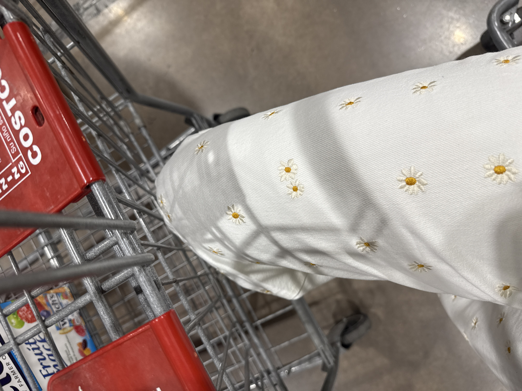 The image shows a person's leg wearing white pants embroidered with daisy patterns inside a Costco shopping cart. The shopping cart also contains a box of "Fruit Sauce" brand fruit snacks. The scene is indoors, presumably within a Costco store, likely located in the city of Puebla, Mexico given the store branding.