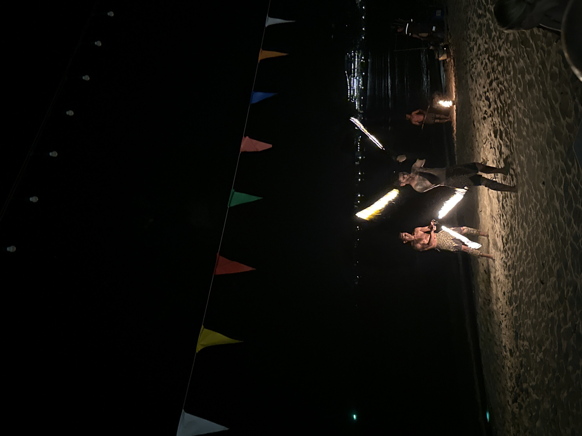 The image depicts a nighttime scene on a sandy beach, likely in Hat Khai Muk, Thailand. Three individuals are performing fire dance with flaming batons near the water's edge. The beach sand appears textured and lit from the fire. In the background, there are decorations including pennant flags, lights and the water. The scene is overall dark, with illumination primarily from the fire.